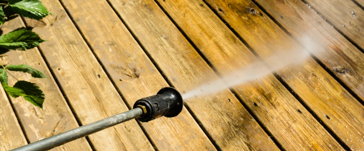 A pressure washer nozzle spraying a powerful stream of water onto a weathered wooden deck, cleaning it.