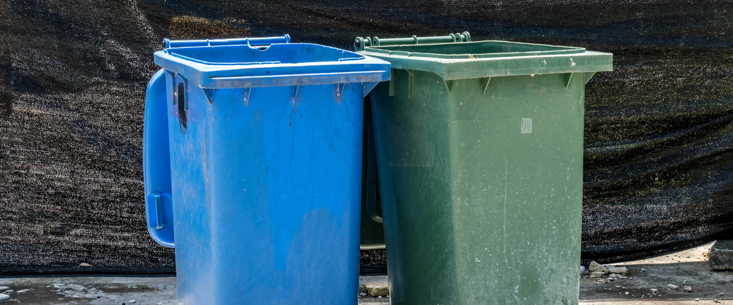 A weathered blue recycling bin stands next to a green waste bin against a dark mesh fence on concrete.