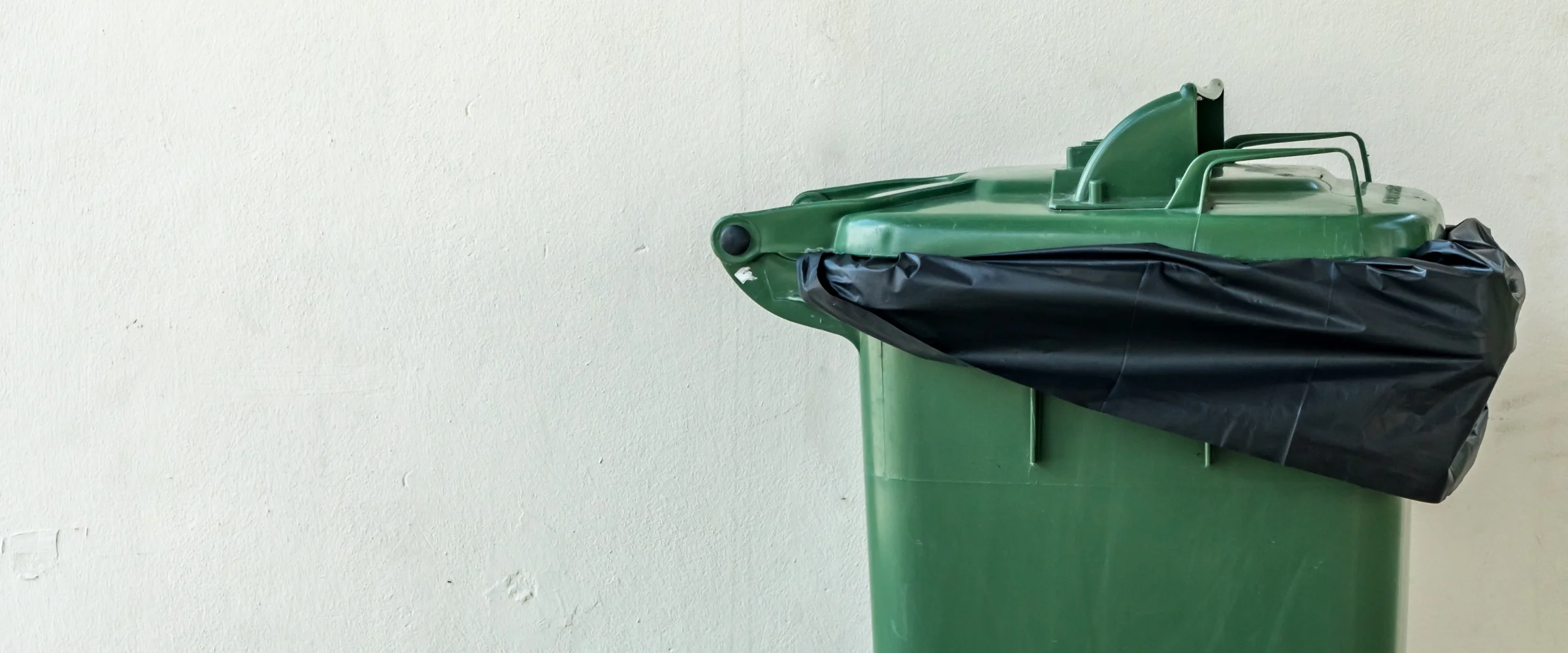 A green garbage bin with a black trash bag overflowing from the top, positioned on the right against a light wall.