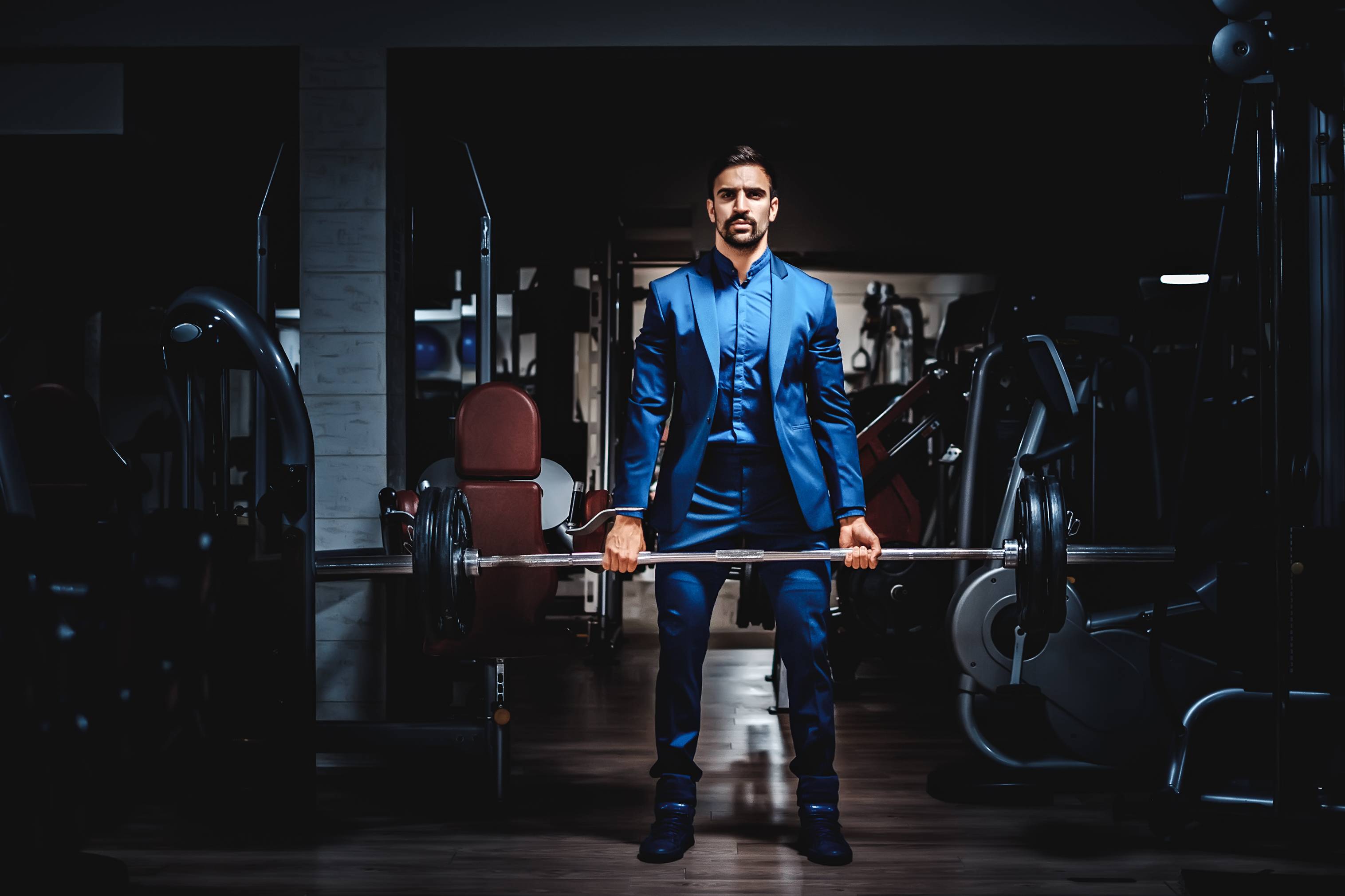 A bodybuilder in a deep blue custom suit that fits his frame. He is holding a bar with weights on it over a bench press.