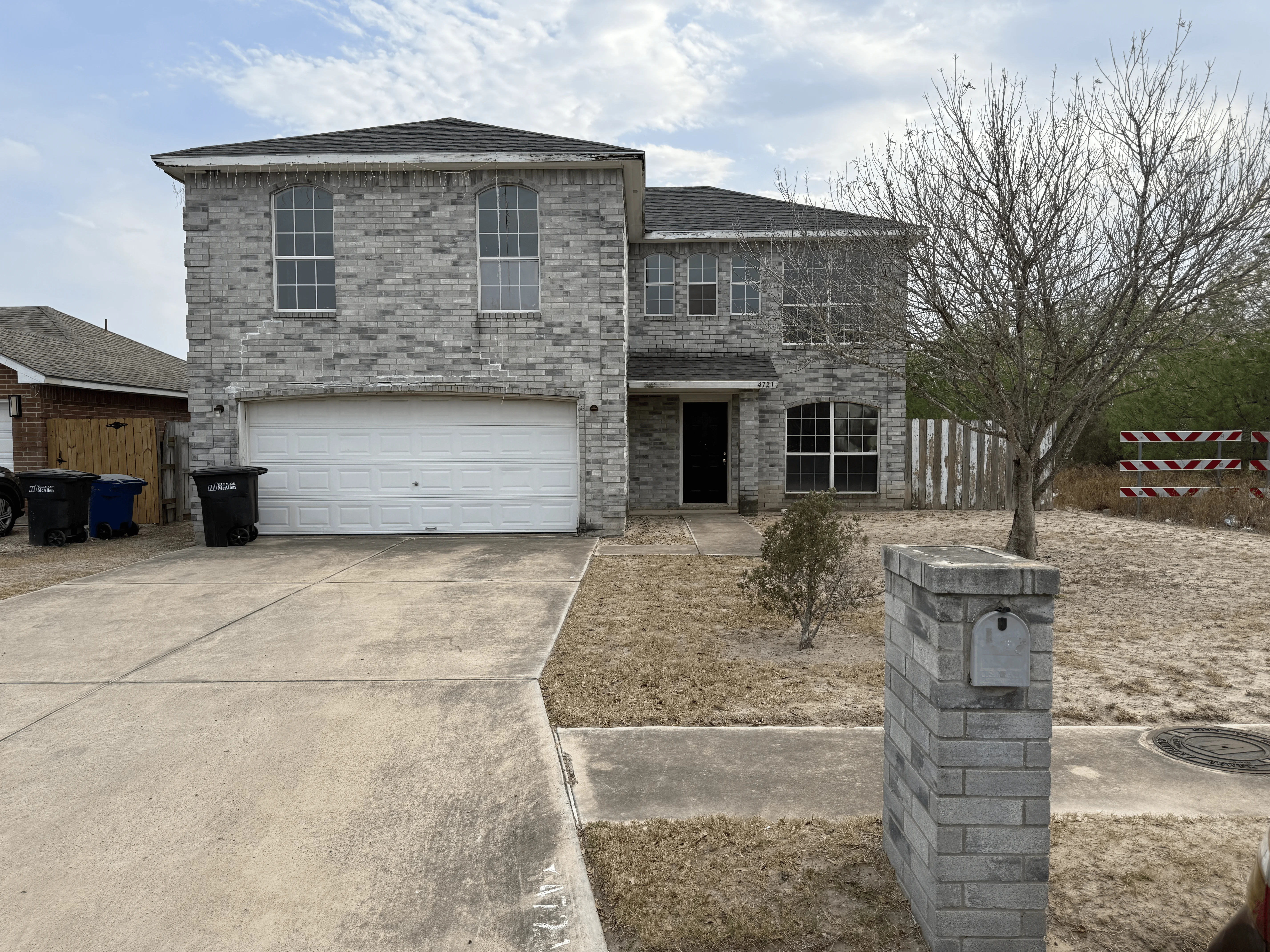 A two-story grey brick house with a white garage door, concrete driveway, and dry front yard.