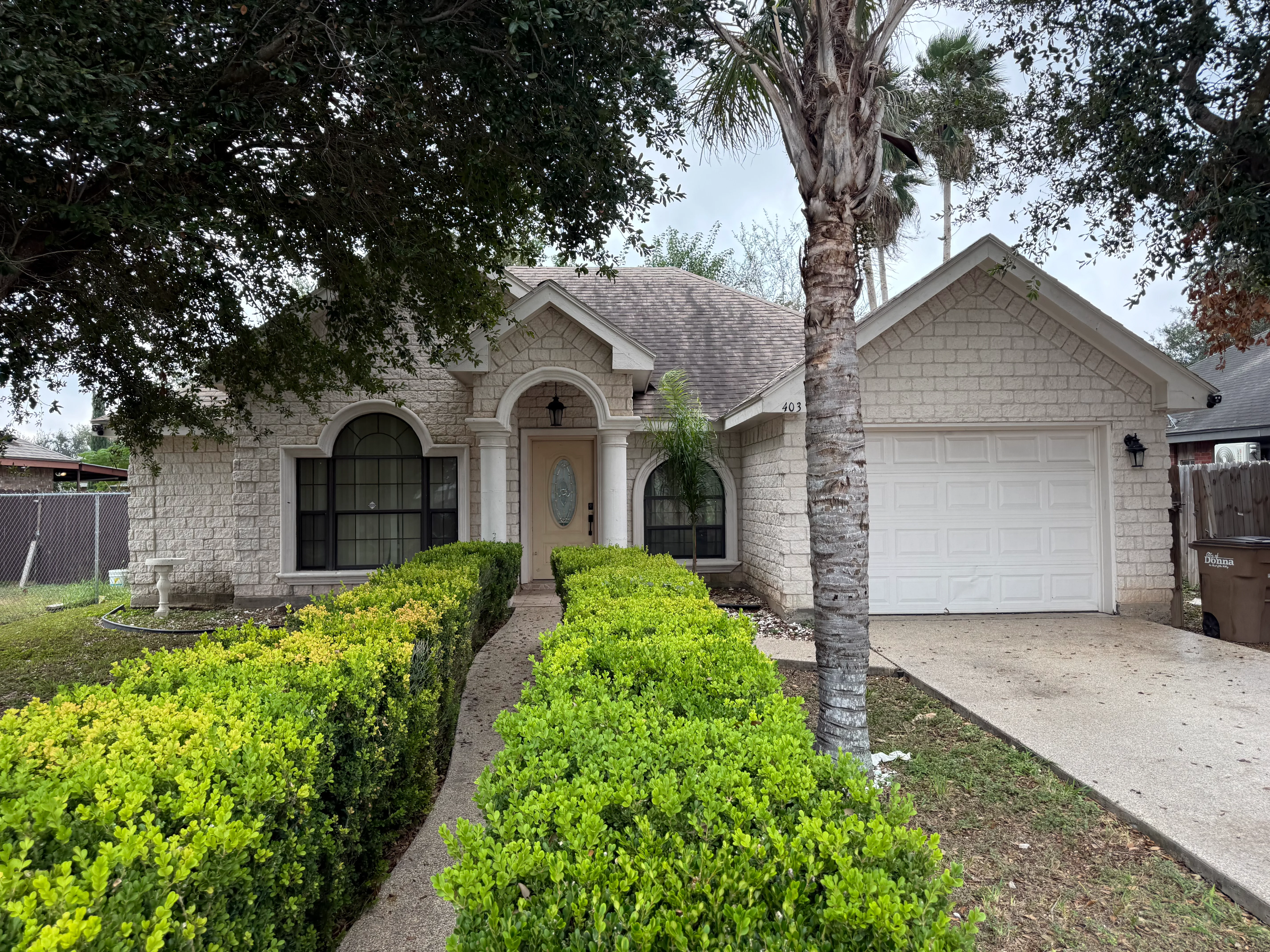 A light-colored stone house with an arched entryway and a paved path leading to it, lined with green bushes.