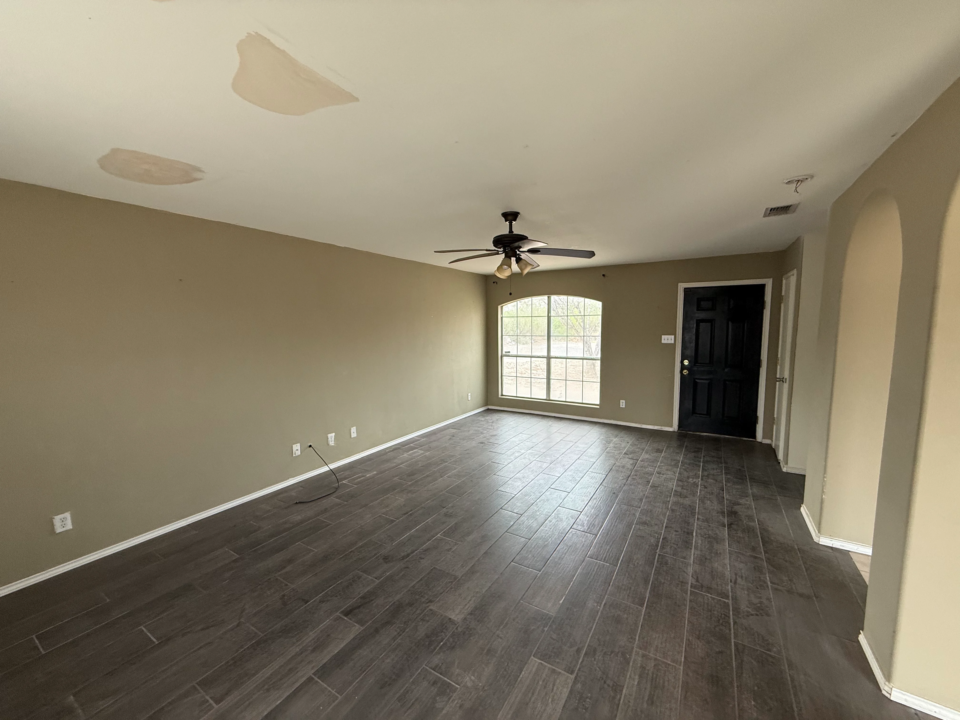 An empty living room with light brown walls, dark gray wood flooring, a ceiling fan, and an arched window.