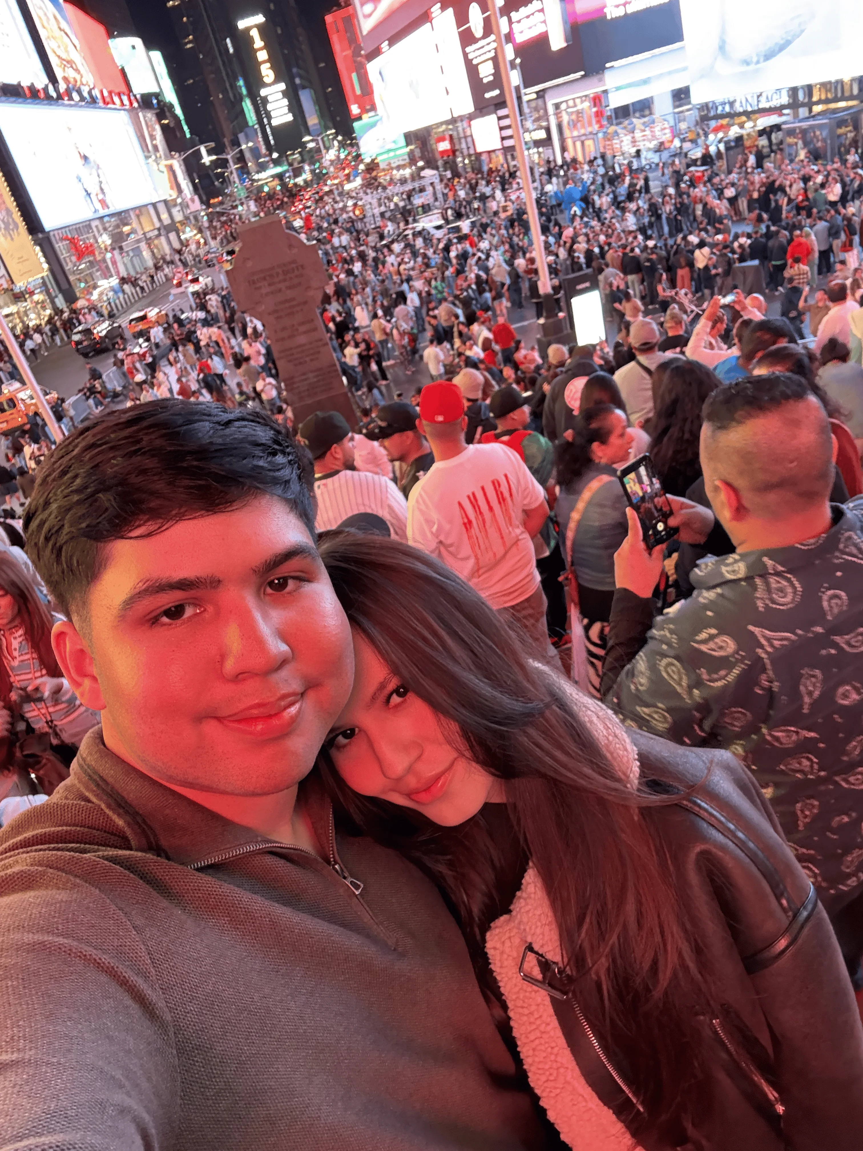 A close-up selfie of a smiling couple in Times Square at night, with a bustling crowd and bright billboards in the background.