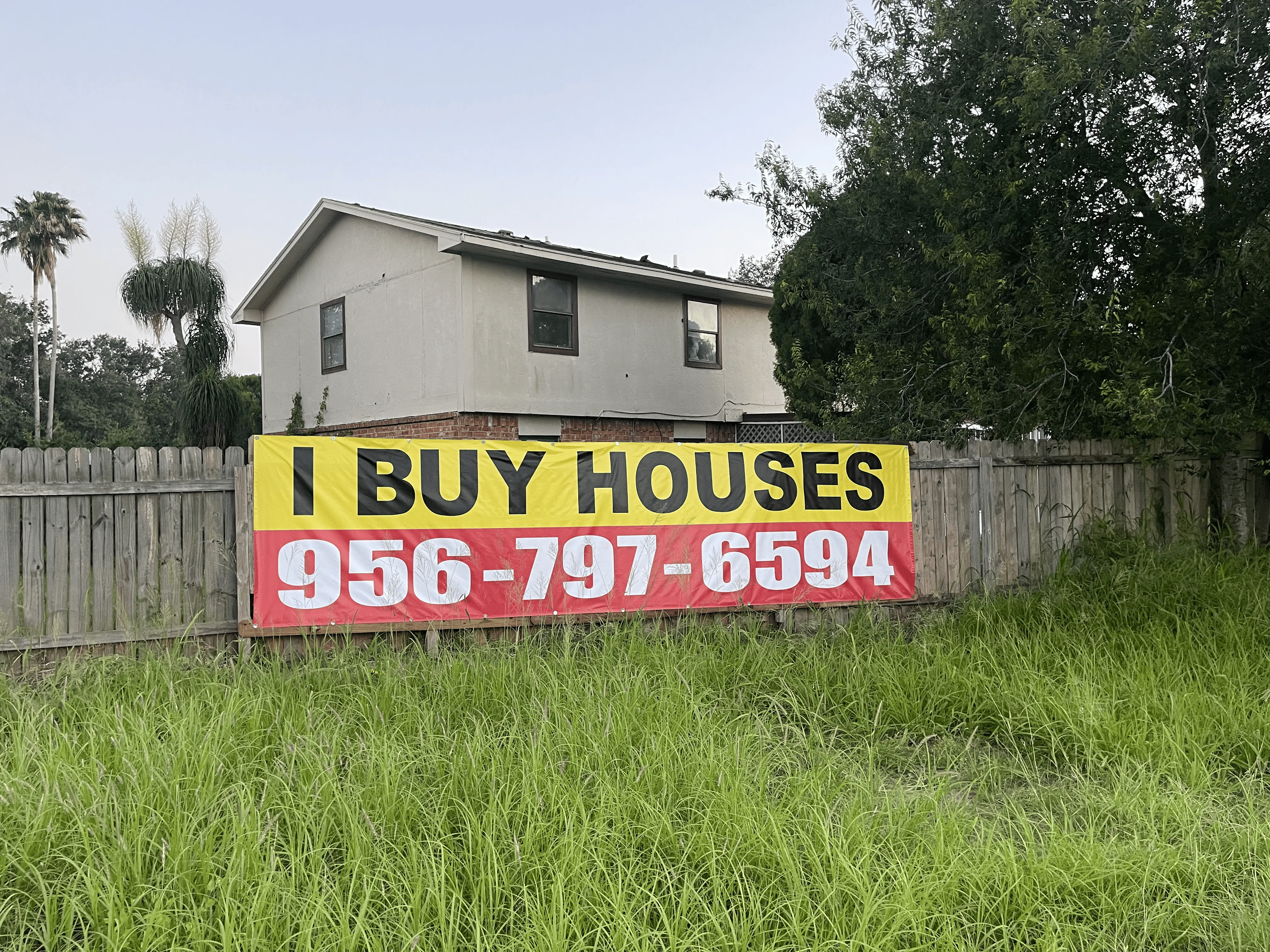 A yellow and red banner reading "I BUY HOUSES" and a phone number hangs on a wooden fence in grassy field.