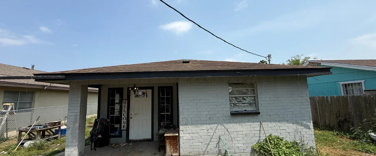 A partially dilapidated house with white brick, a dark roof, and a visible welcome sign by the front door on a sunny day.