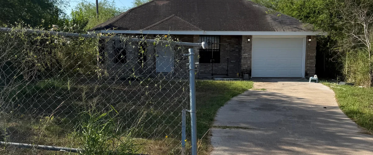 A neglected brick house with a white garage door, viewed through an overgrown chain-link fence and concrete driveway. A neglected brick house with a white garage door, viewed through an overgrown chain-link fence and concrete driveway.