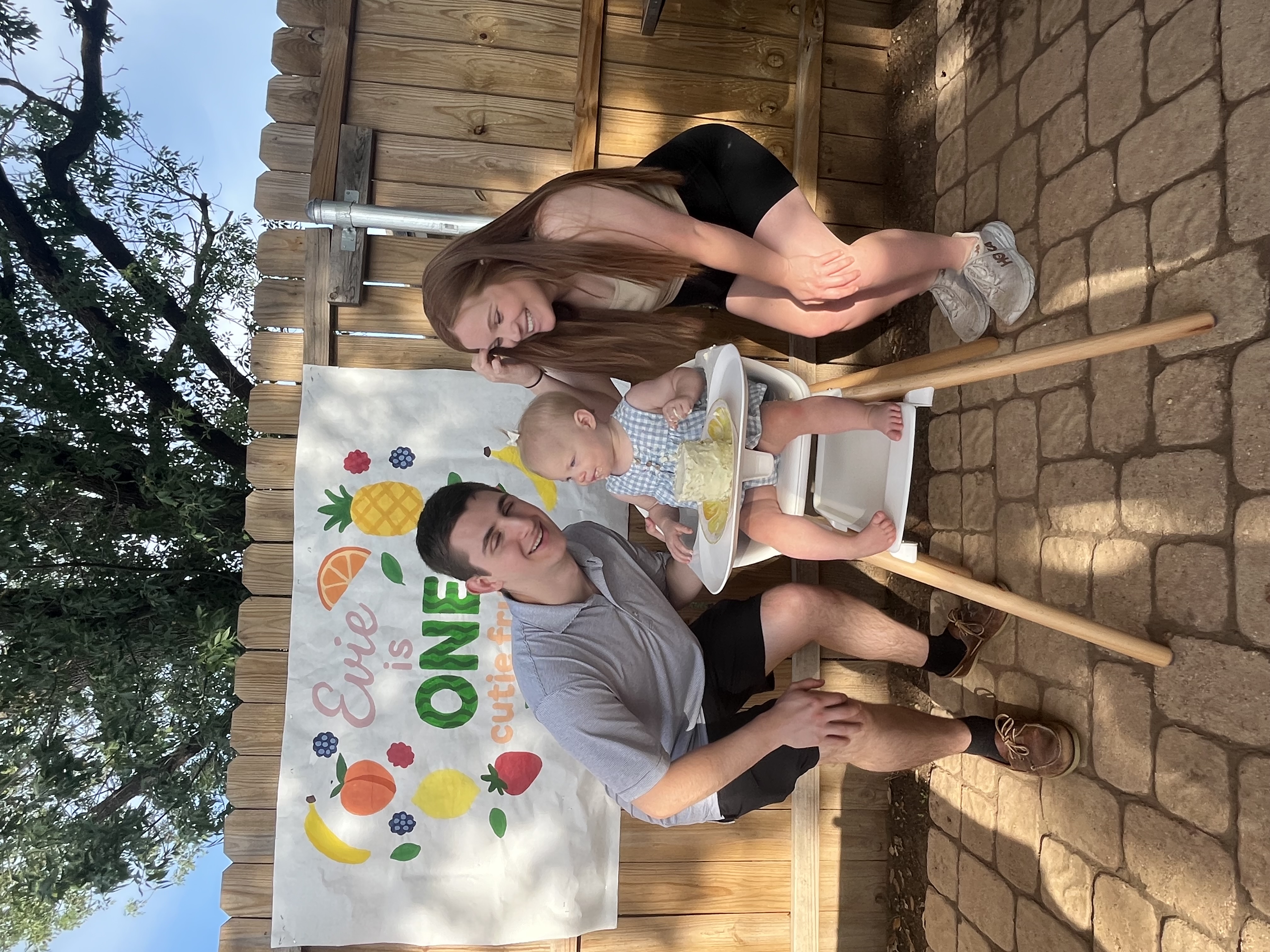 A young family celebrates their daughter's first birthday outdoors. The parents smile and crouch beside their baby, who is seated in a high chair with a small birthday cake. A colorful banner behind them reads "Evie is ONE" with fruit illustrations and the phrase "cutie from the fruitie.