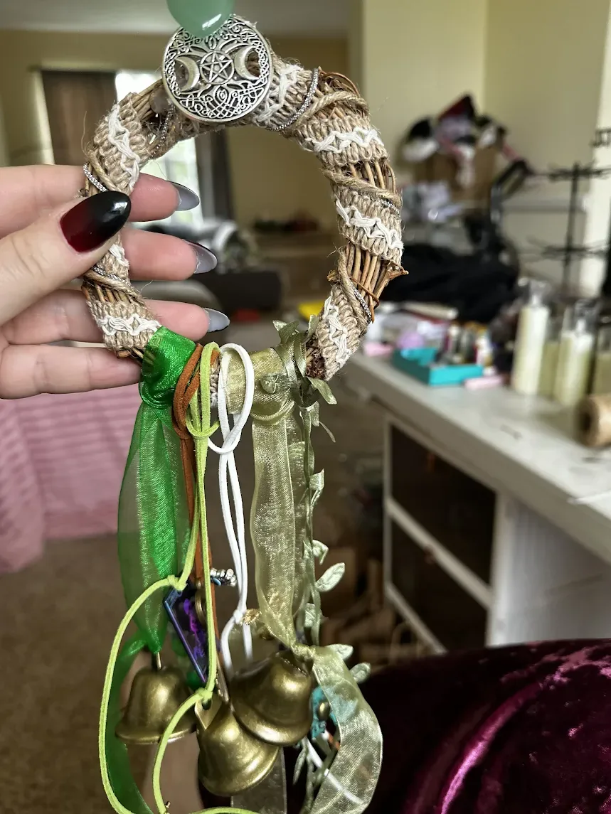 Her hand, with dark red nails, holding a jute-wrapped grapevine bell ring topped with a Triple Moon charm, green ribbons and brass bells hanging below.