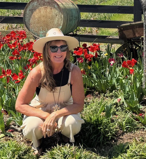 Woman crouches down in a tulip garden, smiling, wearing black tinted sunglasses, a sun hat, and khaki overalls.