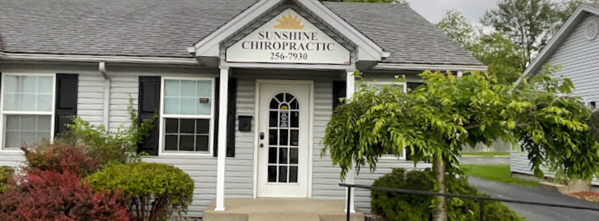 White Chiropractic office with dark blue shutters around windows and a grey shingled roof with a sign featuring a yellow sunshine and the name Sunshine Chiropractic in bold letterng.