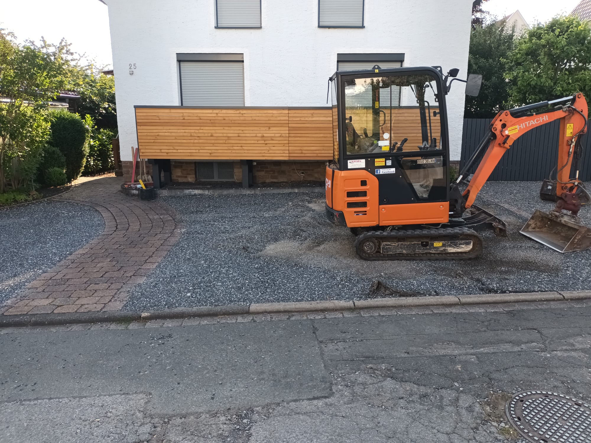 Kompaktbagger auf Schotterfläche in Einfahrt, Vorbereitung des Unterbaus vor Hausfassade