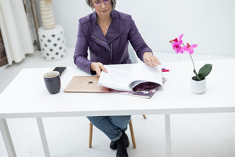 Woman sitting at a desk reviewing an open book beside a laptop, coffee cup, and potted orchid. Woman sitting at a desk reviewing an open book beside a laptop, coffee cup, and potted orchid.
