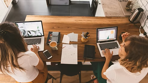 Two women working together on laptops at a shared desk. Two women working together on laptops at a shared desk.