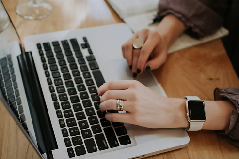 Close-up of a person typing on a laptop at a wooden desk while wearing a smartwatch. Close-up of a person typing on a laptop at a wooden desk while wearing a smartwatch.