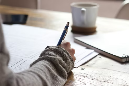 Person writing notes with a pen on paper at a wooden desk with a coffee cup. Person writing notes with a pen on paper at a wooden desk with a coffee cup.