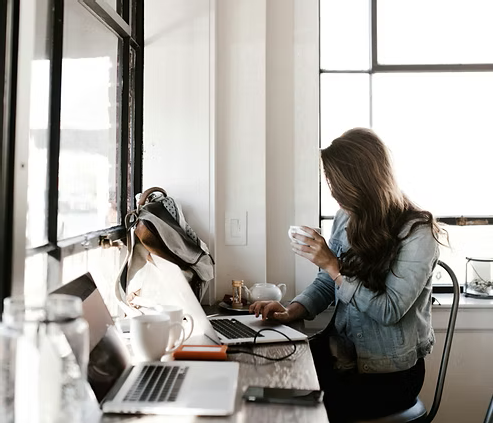 Woman working on a laptop at a bright café or home workspace while holding a cup. Woman working on a laptop at a bright café or home workspace while holding a cup.