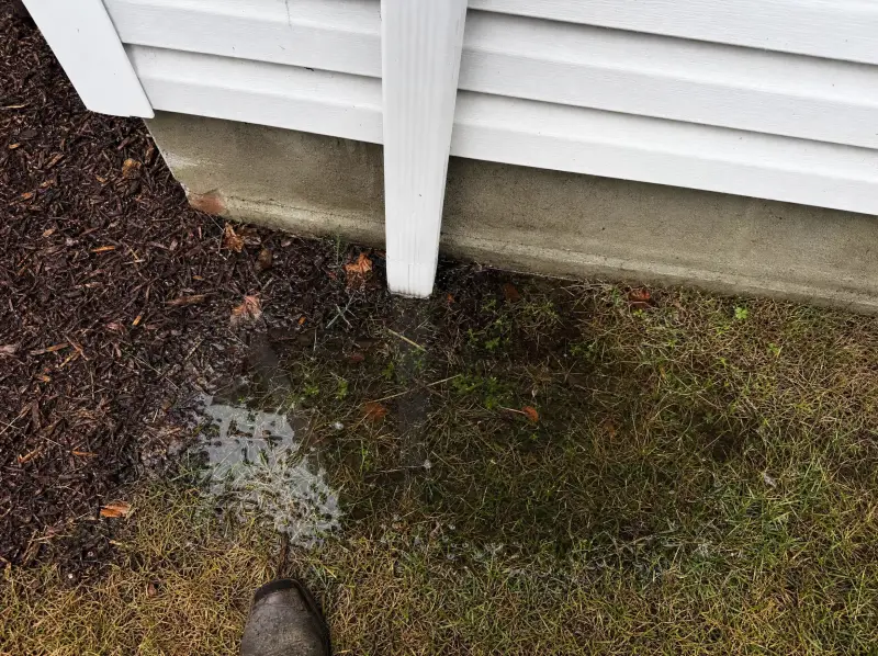 Illustration: a downspout ending flush at the foundation with water standing in the saturated ground beside it.