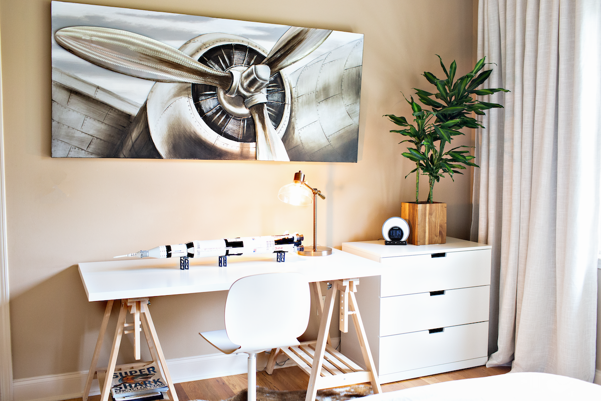 White wooden desk with matching chair, green plant, and wall art of a plane.