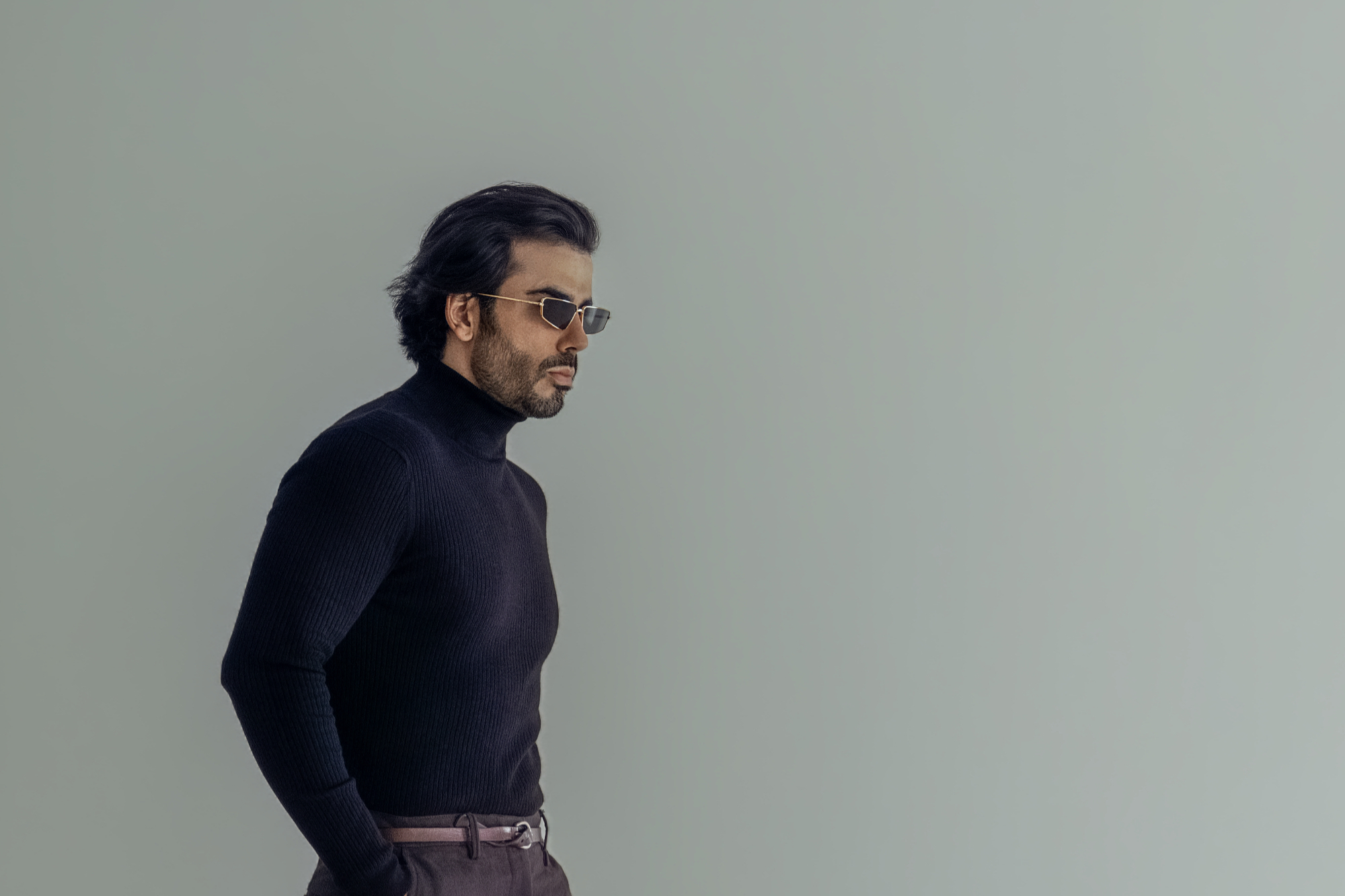 Man in a black turtleneck and gold rimmed sunglasses against a sage wall