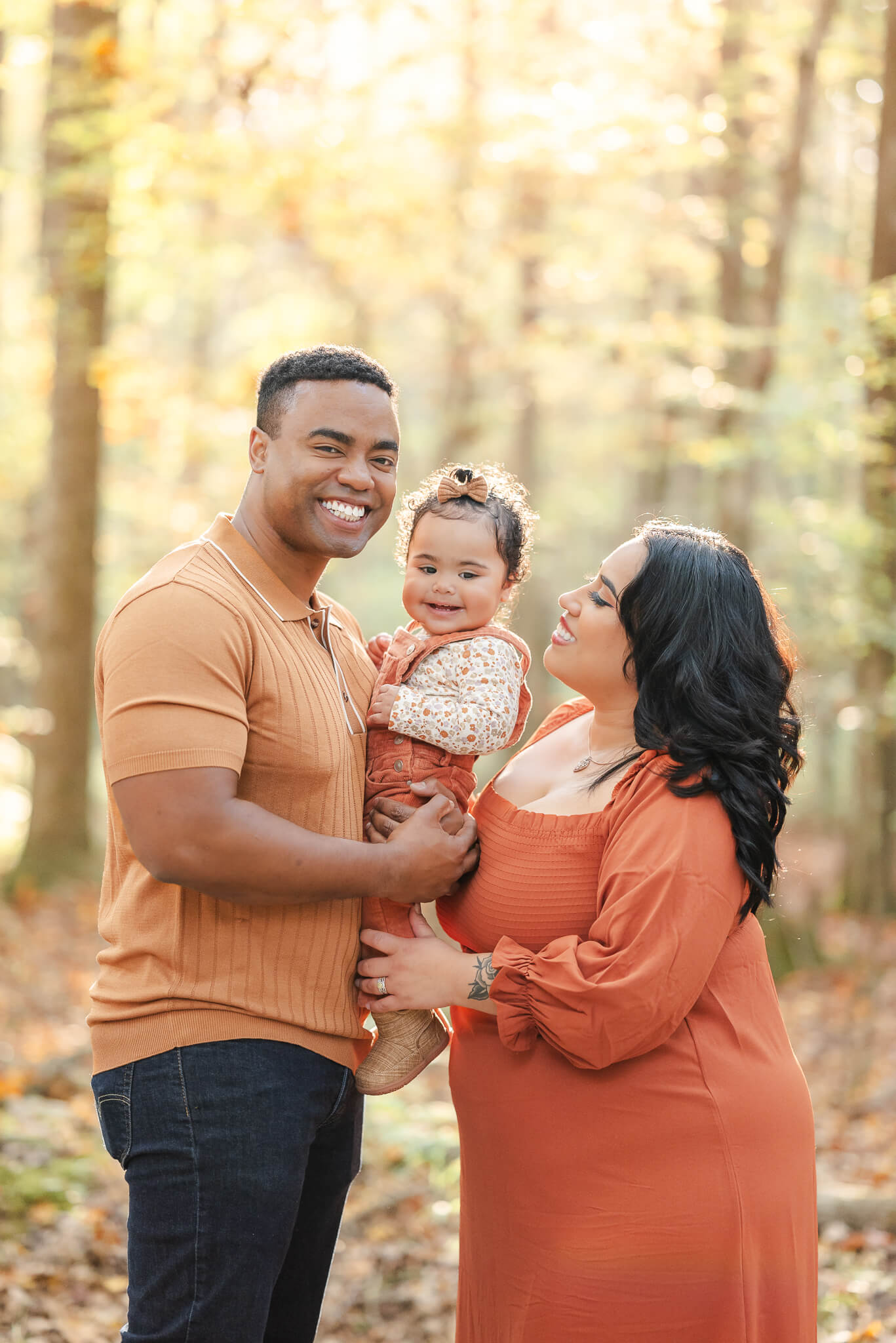 A family of 3, stands in a forest with the sun shining between the trees behind them. These are fall family portraits, captured by Virginia Beach photographer, Justine Renee Photography. The family is dressed in oranges. Mom is in a dress, dad, in dark jeans and collared shirt. The toddler has on orange overalls with a matching bow in her hair.