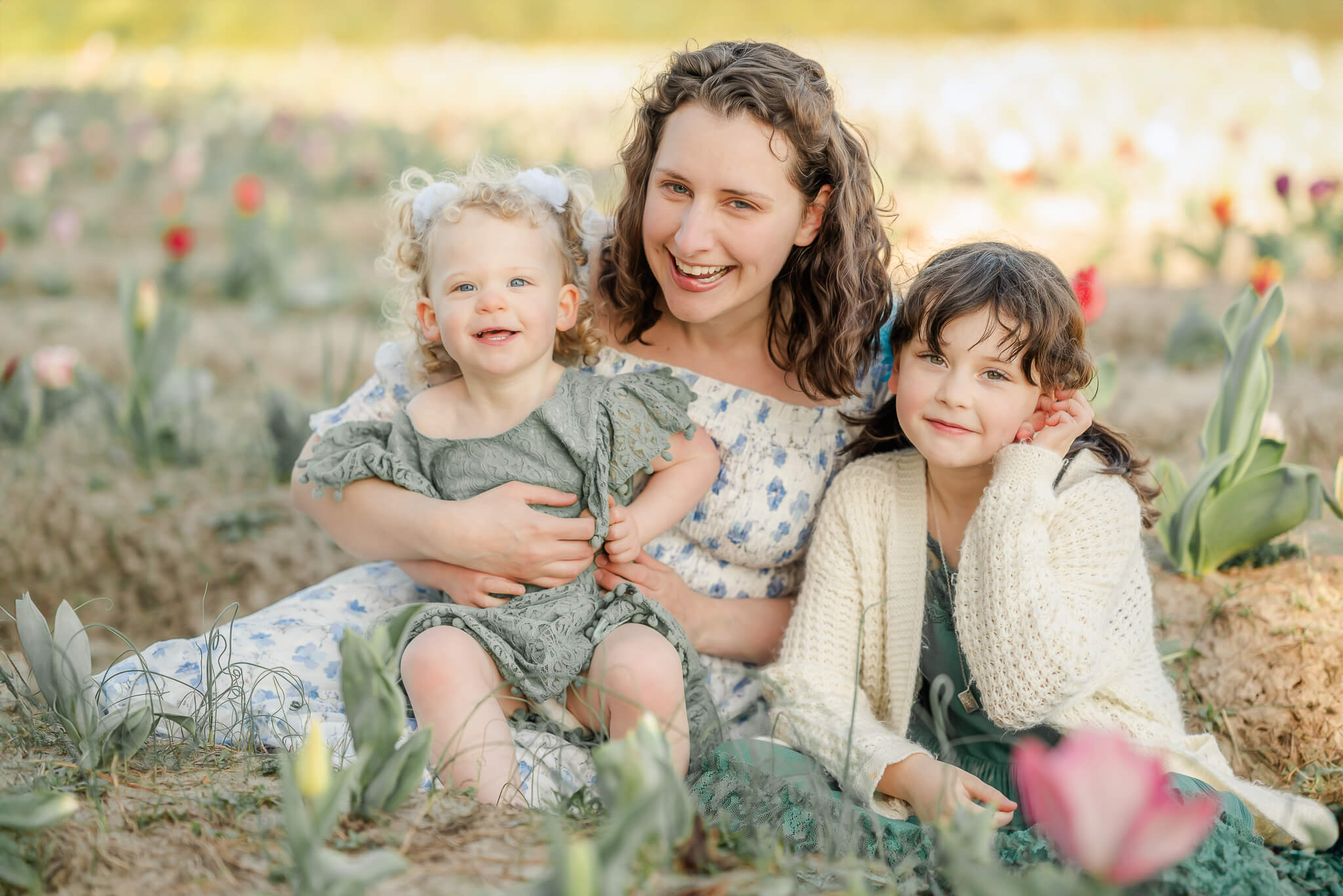 Image of the photographer at Justine Renee Photography. She is sitting in a tulip field laughing with her two young daughters. They are all dress for spring in green and blue dresses.