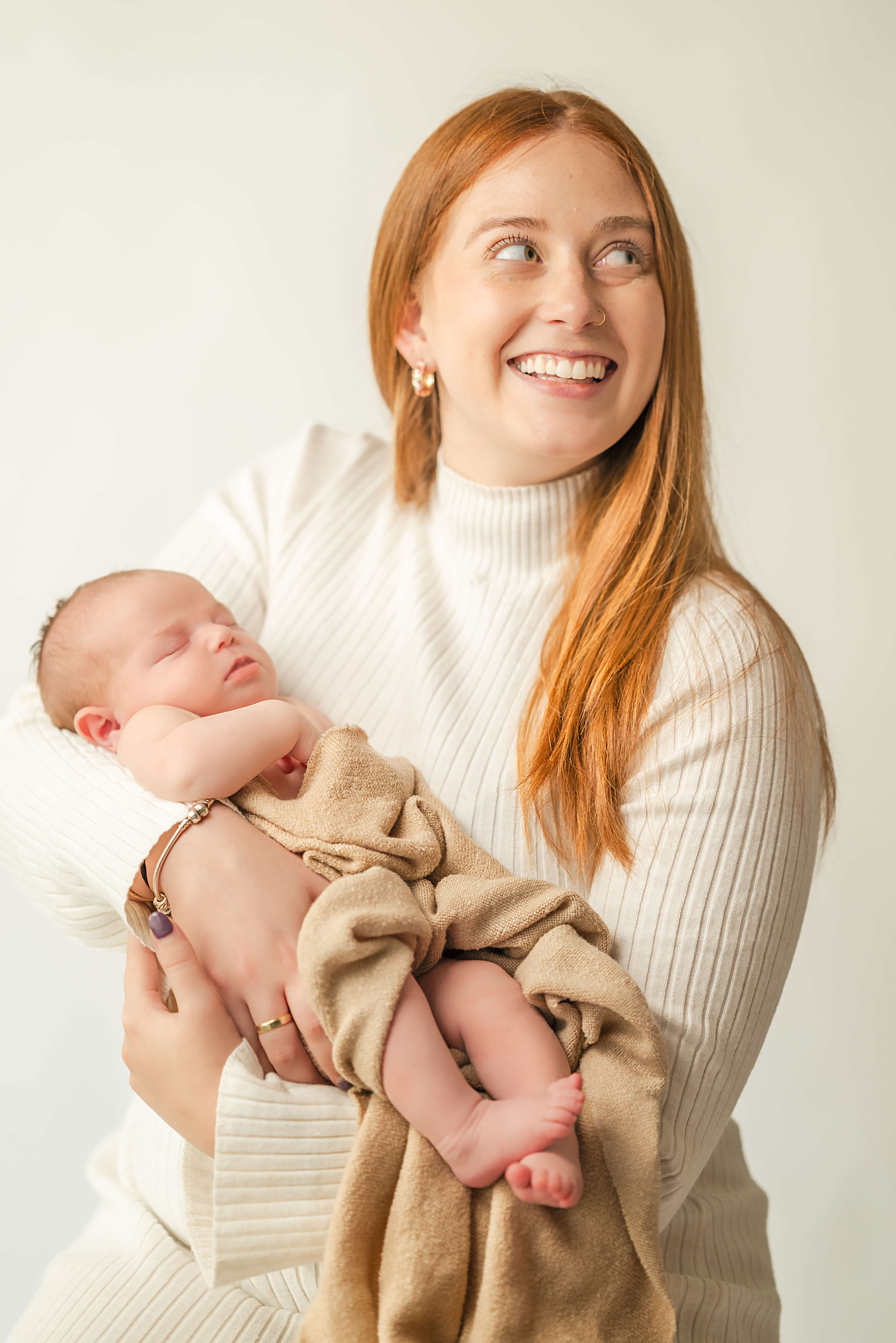 A young mother, holding her newborn baby, captured by Virginia Beach photographer, Justine Renee Photography. The mother, who has long red hair, is wearing an off-white sweater dress and smiling while looking at her husband, who is out of frame. The baby is asleep and wrapped in a light brown blanket.