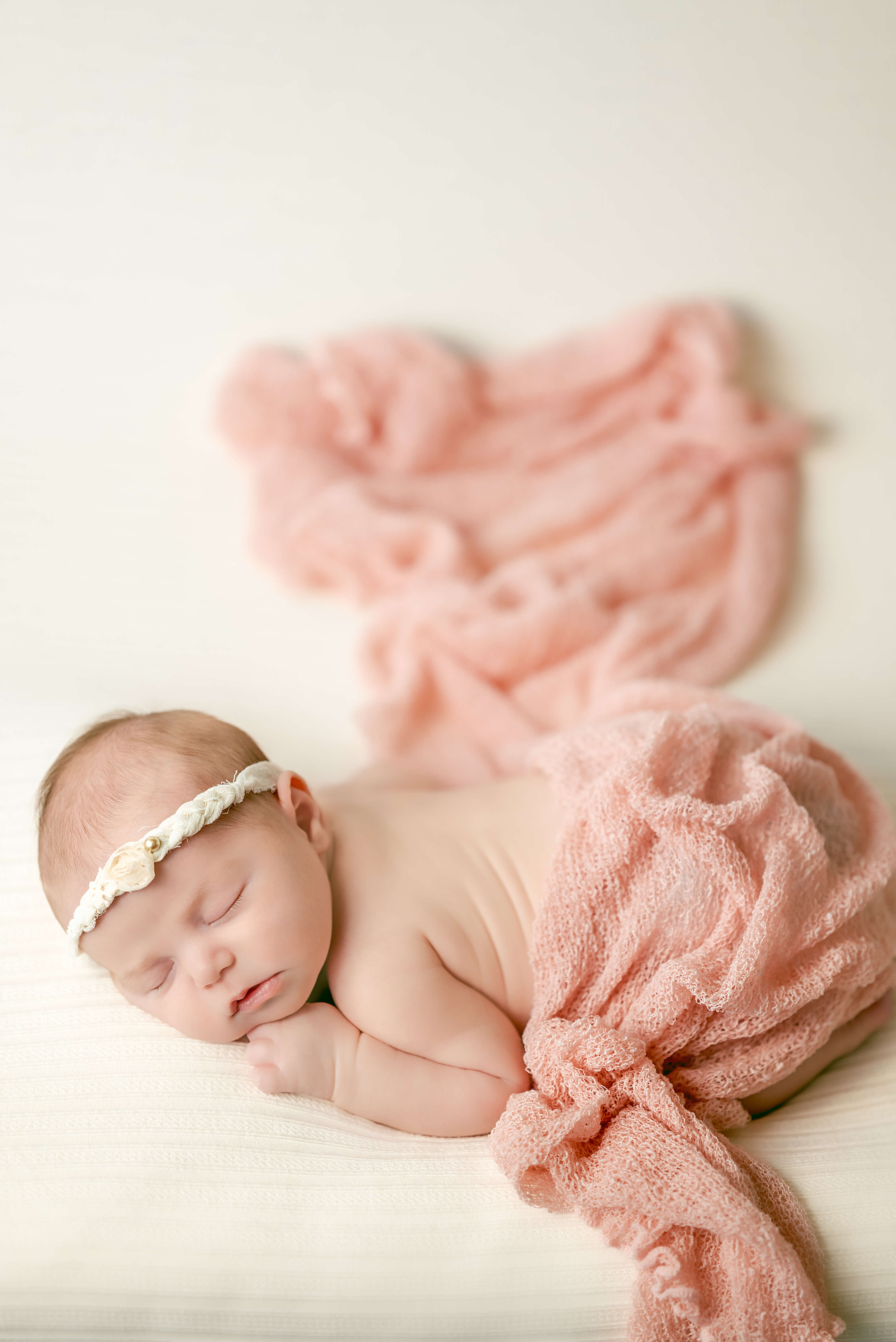 A newborn baby sleeps peacefully on a white blanket. She is wearing a white headband with a small flower on it and draped in a pink blanket. Image captured by Virginia Beach newborn photographer, Justine Renee Photography.