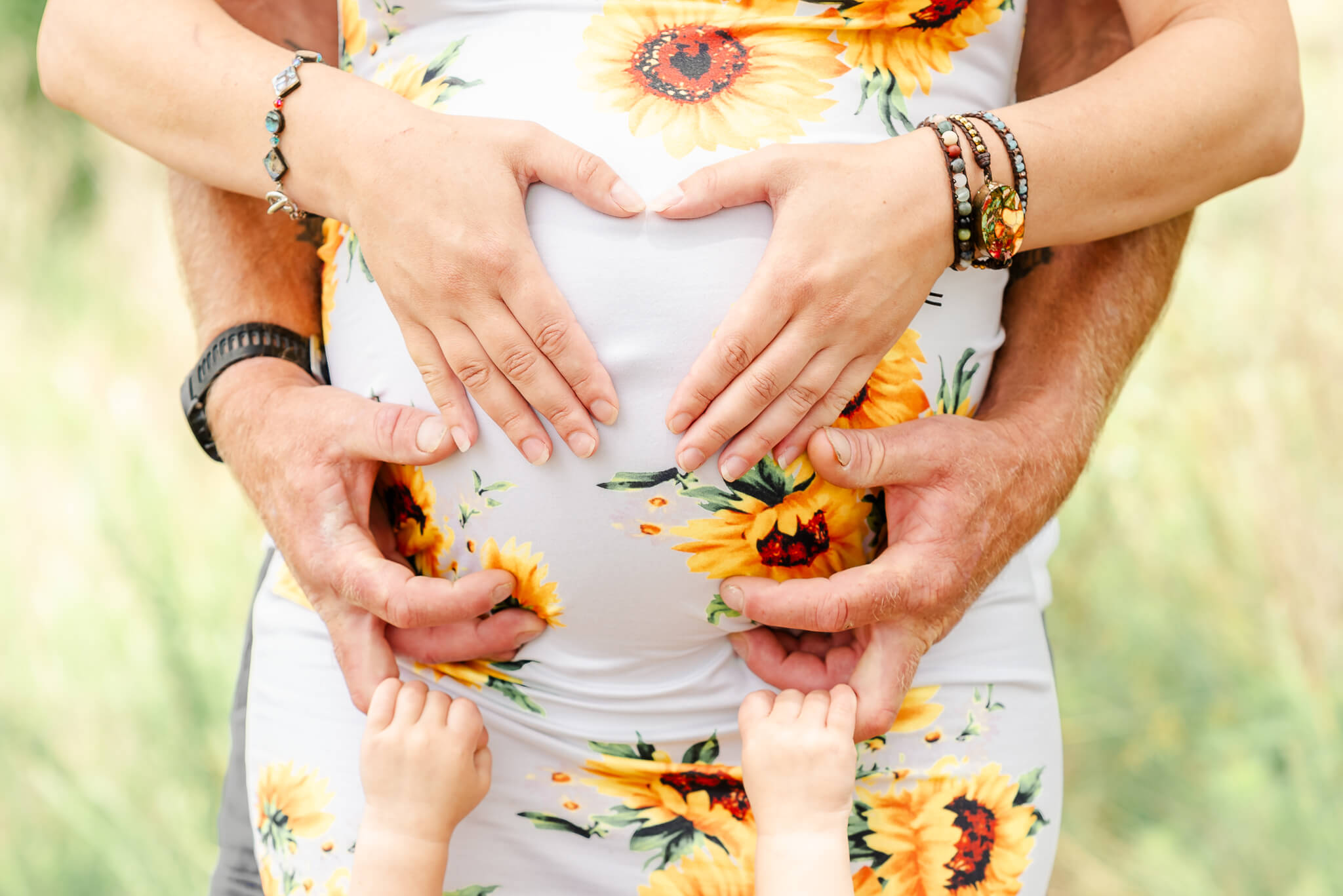 An expectant mother wears a white dress with sunflowers on it. She is making a heart with her hands on her belly. Her partner stands behind her and her toddler in front. The toddler holds her father's hands just below her mothers belly.