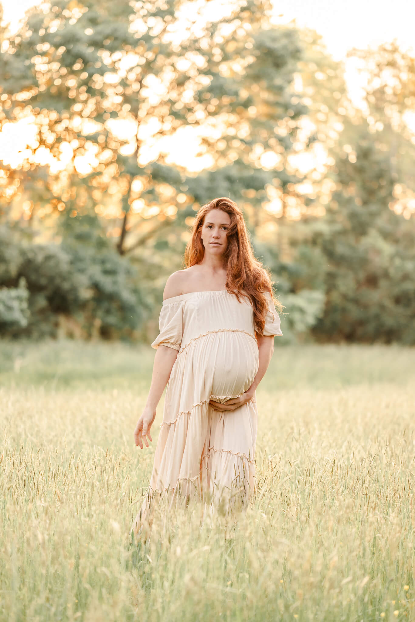 An expectant mother walks through a grassy field while cradling her belly. The sun is shining through the trees behind her and the loose off-white dress compliments her red hair. Image captured by Virginia Beach photographer, Justine Renee Photography.