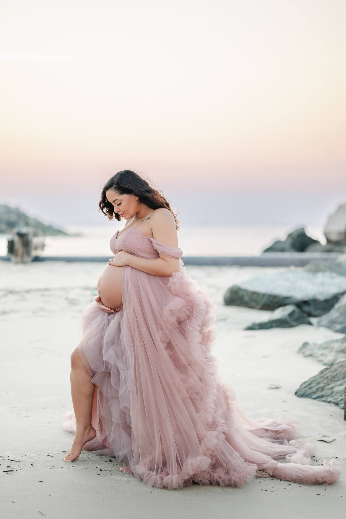 An expectant mother looks down at her growing belly while wearing a pink tulle gown. Her belly is peeking out and her knee is popped. She is standing on the beach. The sun is setting, creating a soft purple and pink sky behind her. Image captured by Virginia Beach photographer, Justine Renee Photography.