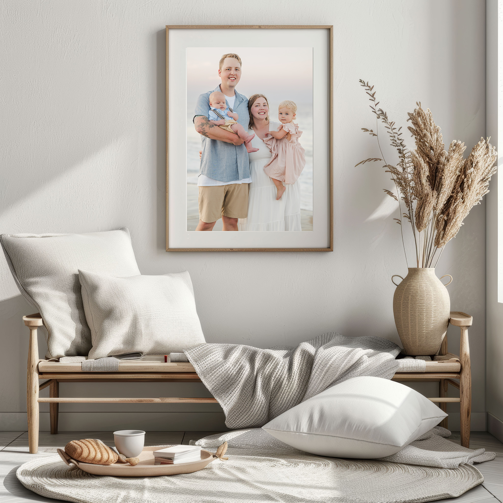 An example of a photograph printed large for the wall. The photo features a young family on the beach at sunset. The toddler girl and baby boy are held in their parents' arms. The photo hangs on a white wall above a wooden bench, decorated with white pillows, a soft gray blanket, and pampas grass in a vase. Large printed portraits can be purchased through Virginia Beach photographer, Justine Renee Photography