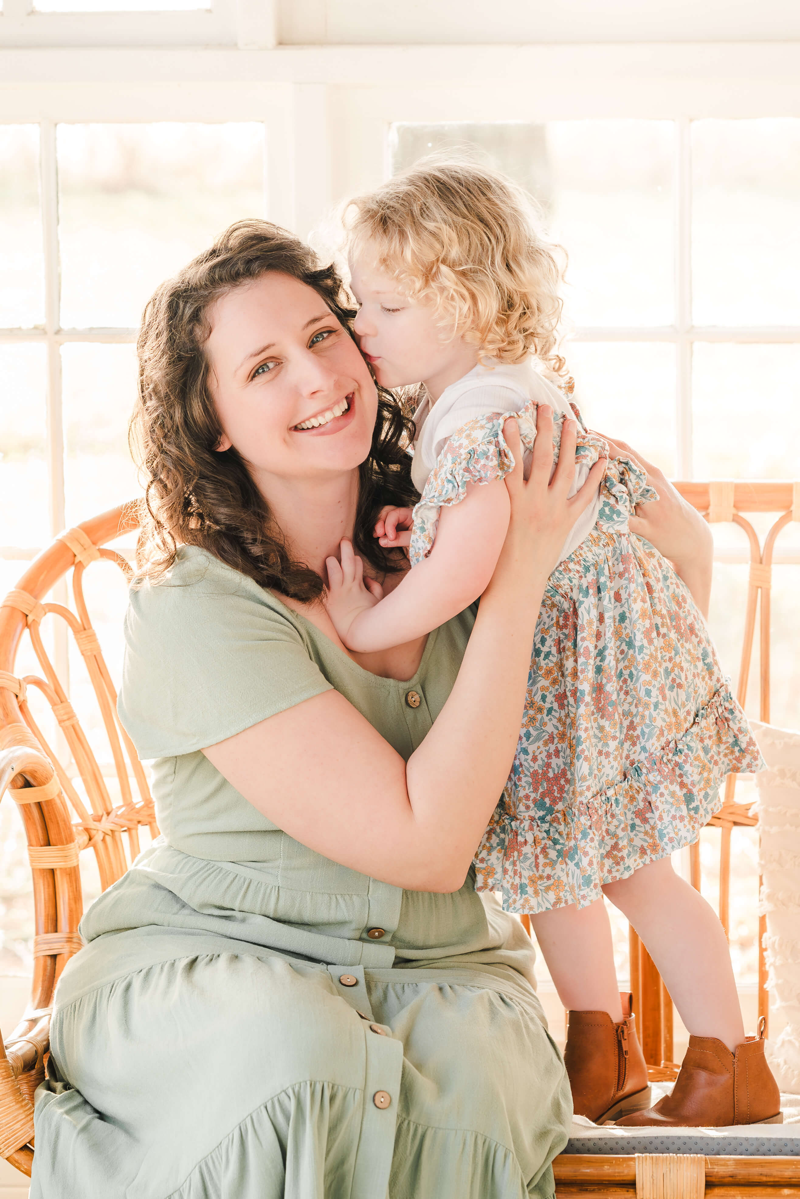 Justine, the photographer at Justine Renee Photography, is sitting on a bench in a sage green dress. Her young daughter, wearing a floral dress and brown boots, stands next to her and gives her a kiss on the cheek.