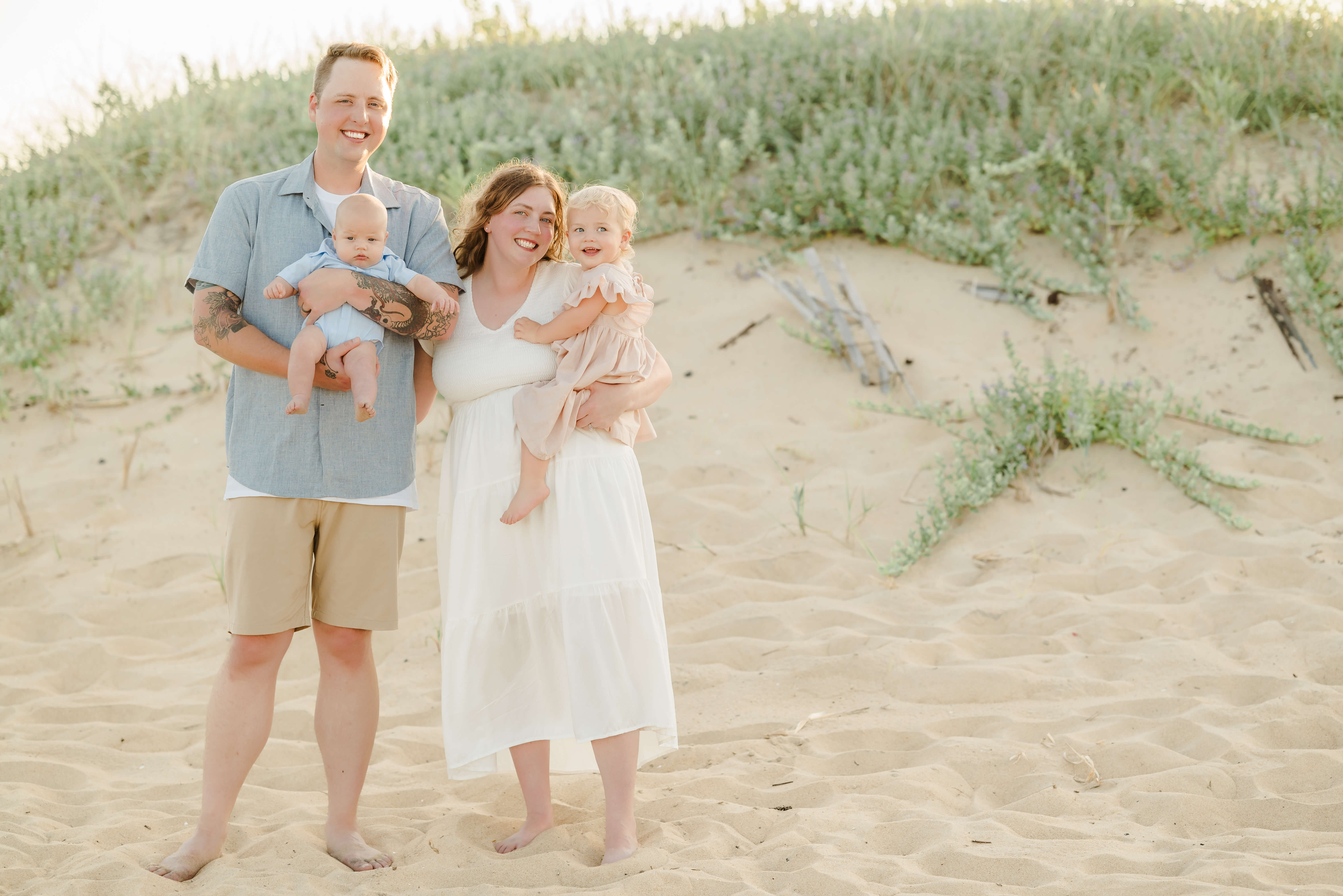 A family of four stands in front of the dunes. The father holds the infant son. Both are dressed in button down blue shirt. The mom, in a white dress, holds the toddler girl, who is in a pink dress.