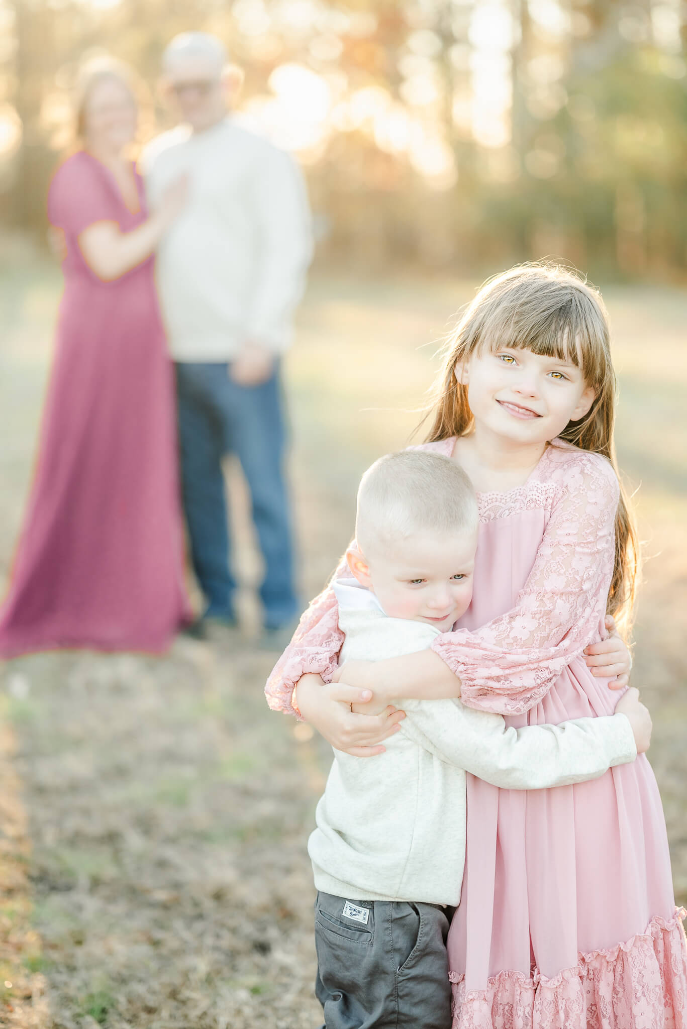 A family portrait captured by Hampton Roads Photographer, Justine Renee Photography. The photo features a brother and sister hugging in the top right corner. Behind them, to the left, the parents stand arm in arm with the sun shining in the trees behind them.