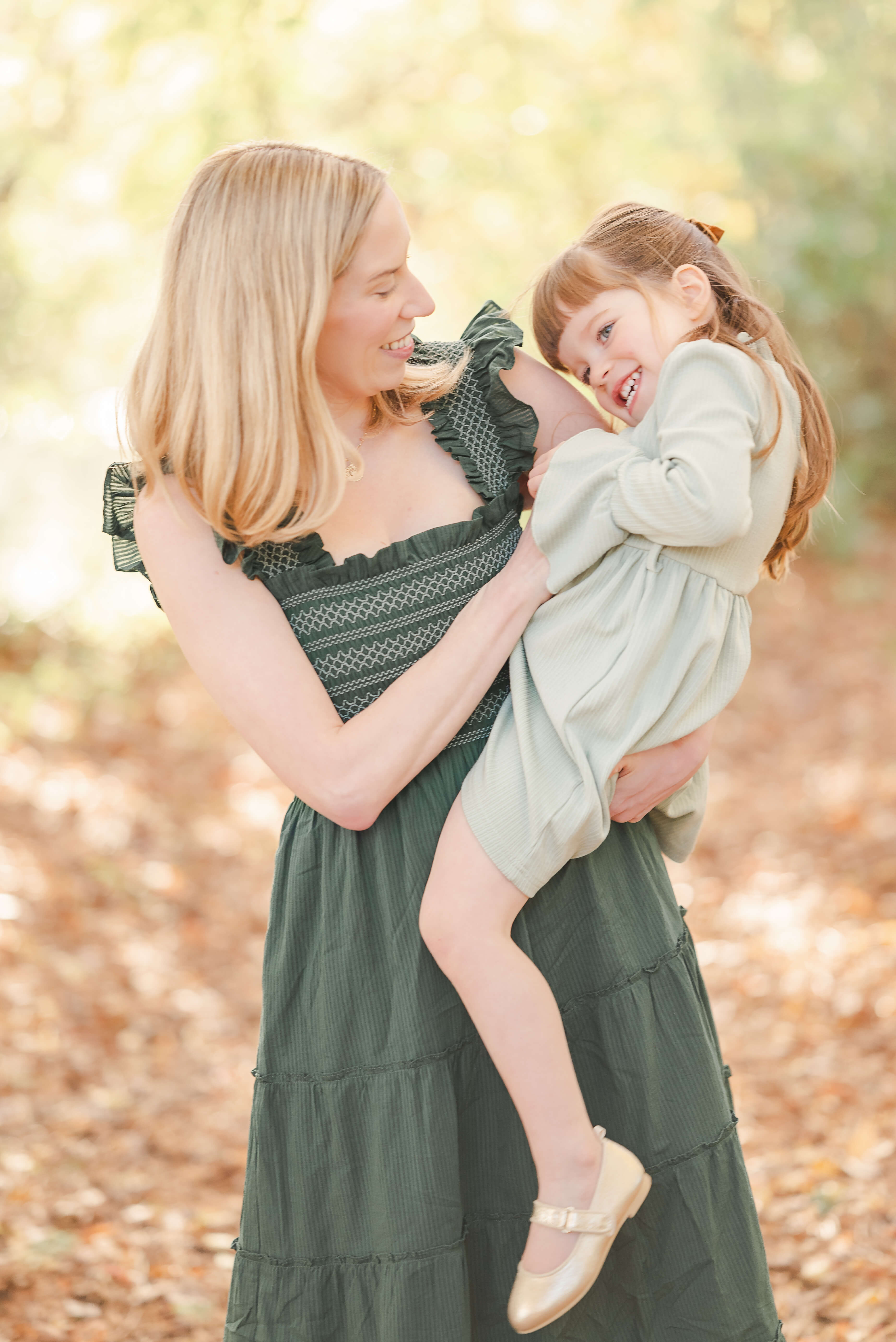 A mother with blond hair, wearing a green dress, tickles her toddler who is on her hip. The little girl, who is in a lighter green dress is laughing.
