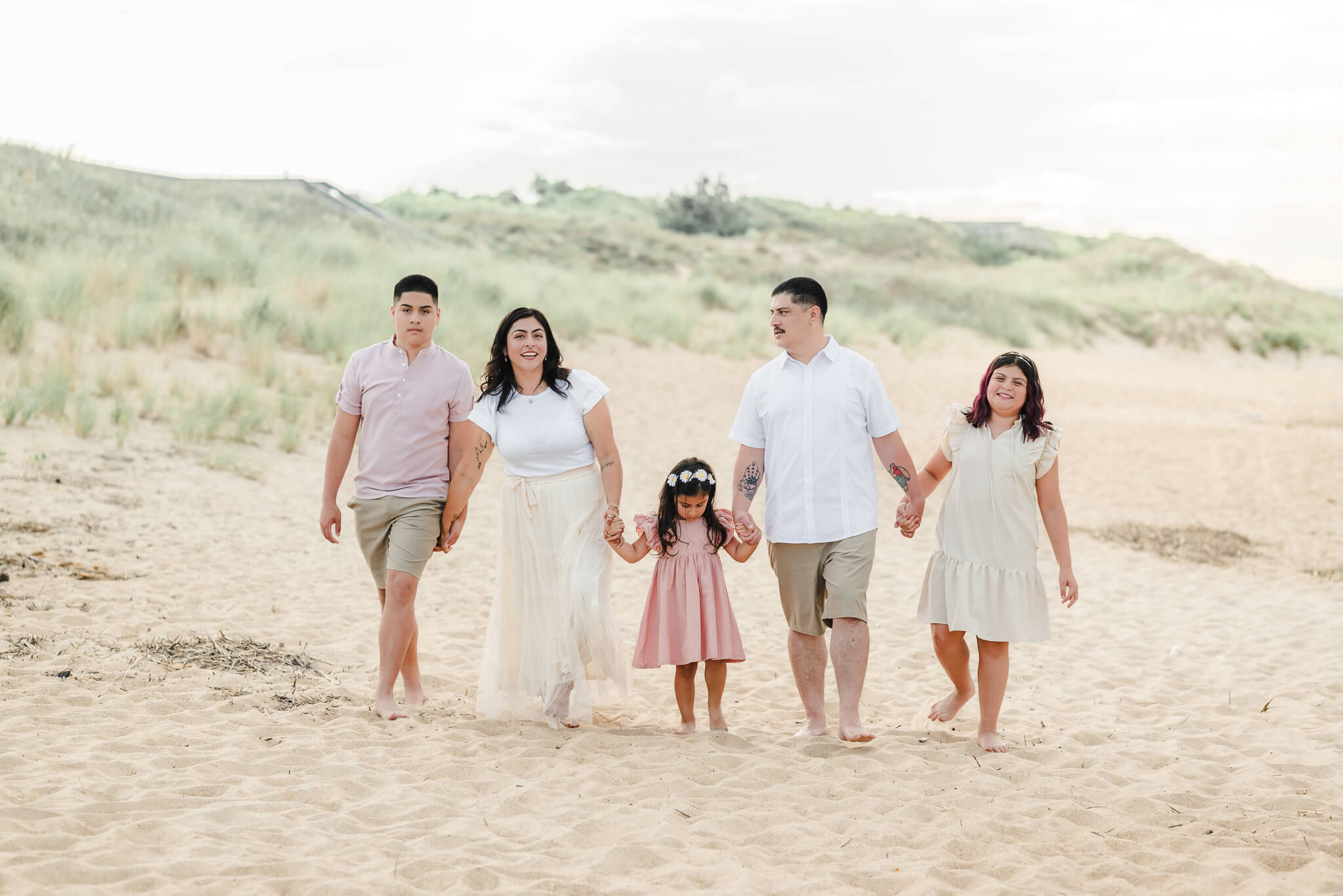 A family of 5 walks along the beach. The sand duns can be seen behind them. The family, who all have black hair, wears a mix of whites, tans, and pinks. Image taken by Hampton Roads photographer, Justine Renee Photography.