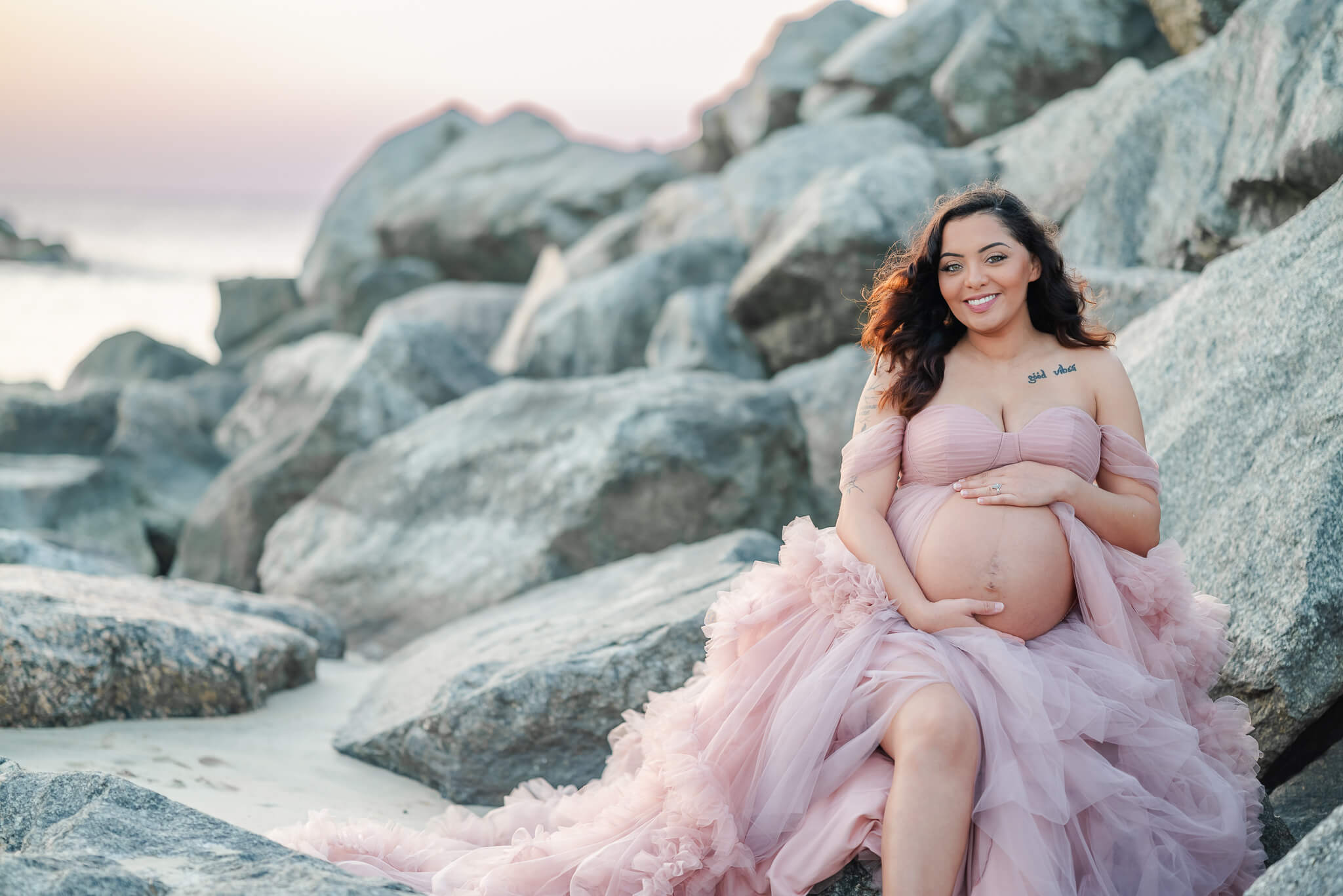 A mama-to-be, in a pink tulle dress, holds her exposed belly and smiles down at it. She is on the beach with the setting sun lighting up the sky in purple, blue, pink, and orange.