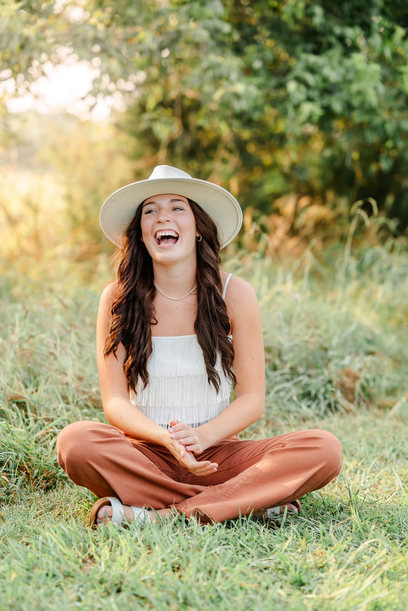 A high school senior, wearing a white top and hat, and brown pants, sits in the grass. The sun is shining through the trees behind her. She is laughing out loud at a prompt from Moyock senior portrait photographer, Justine Renee Photography.