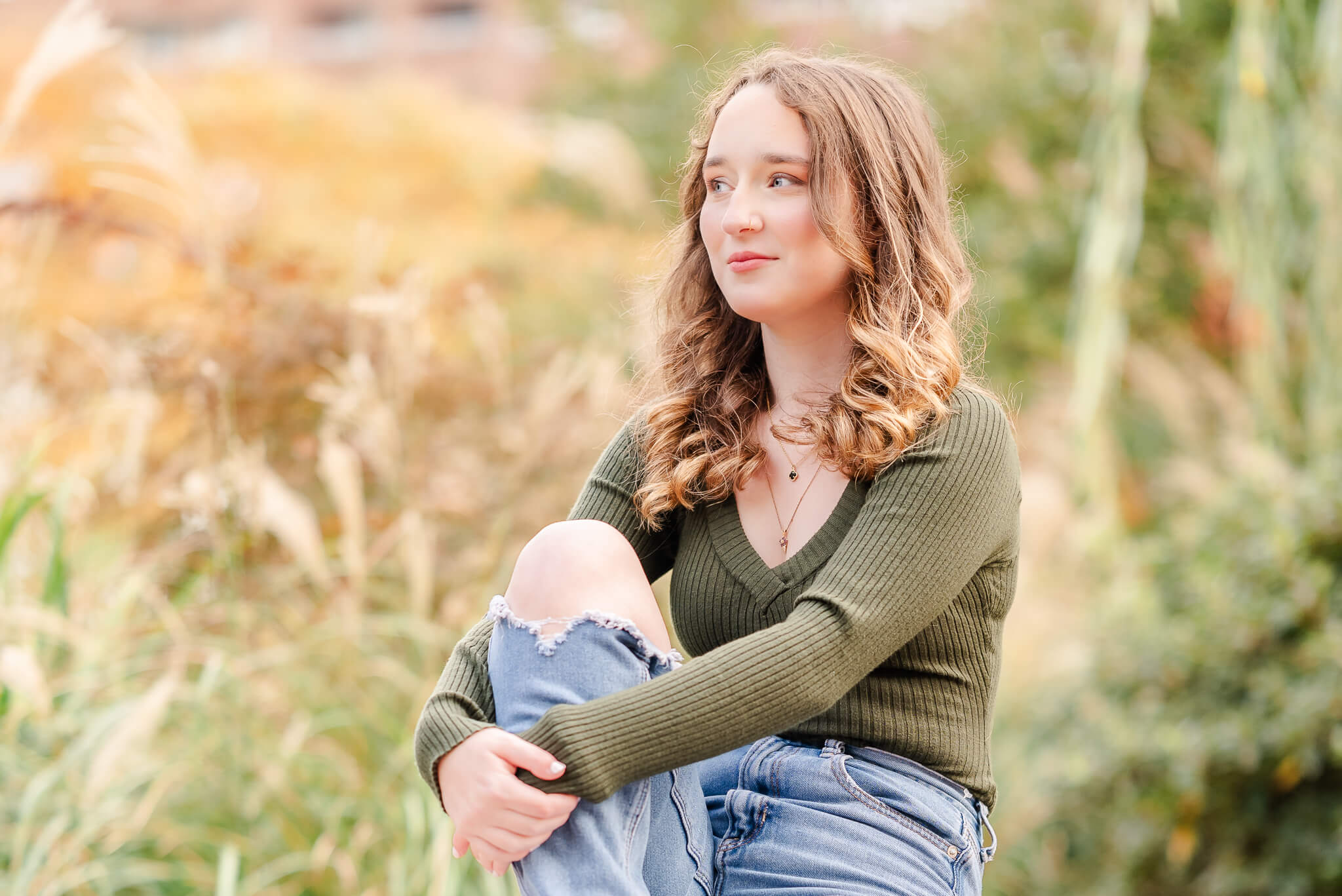 A high school senior poses for her senior portraits with Justine Renee Photography. She is wearing a long-sleeve green shirt and jeans with a tear at the knee. Behind her there is pampas grass and other greenery.