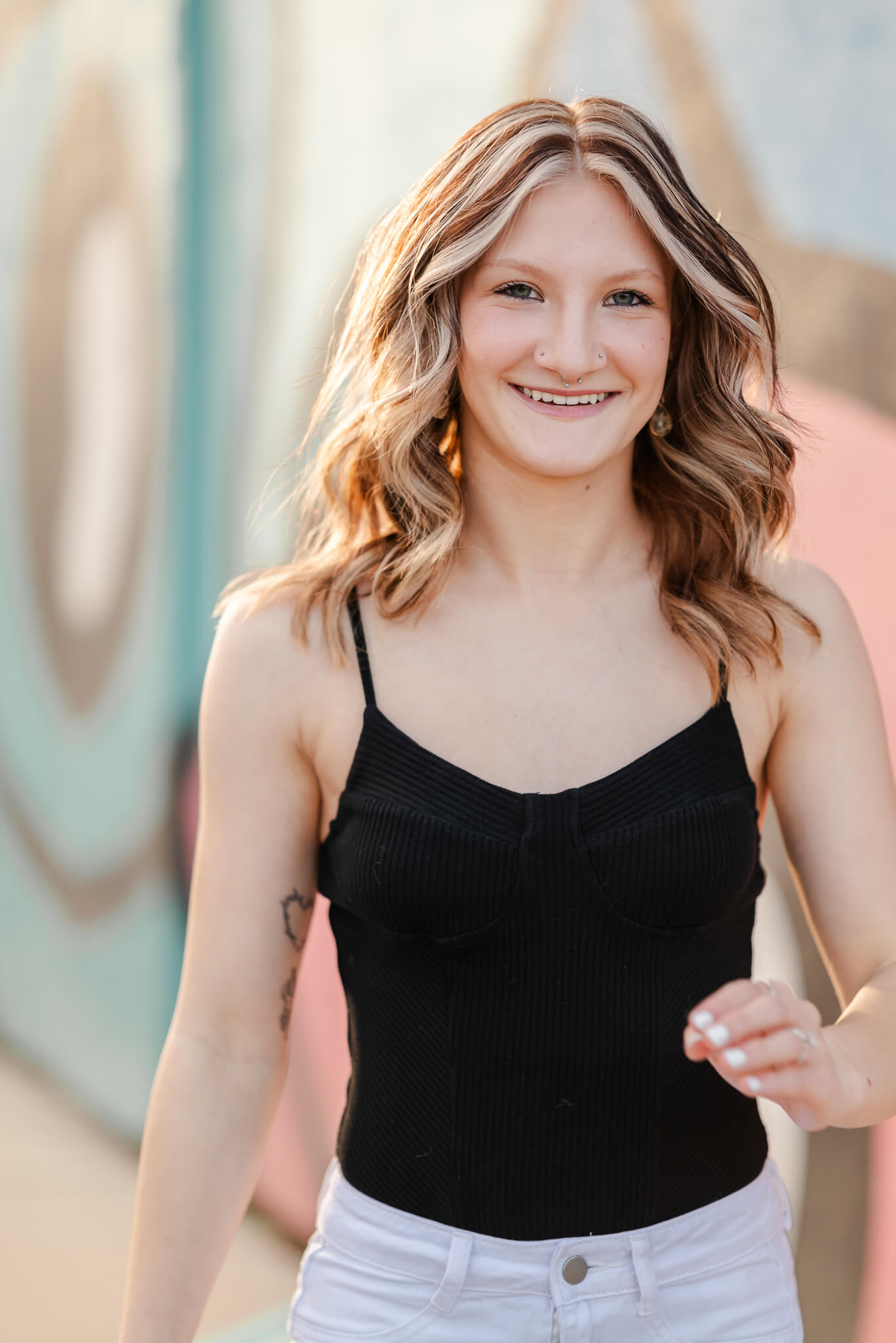 A high school senior, wearing a black tank and white skirt walks with a big smile on her face. A colorful, sun-soaked wall is behind her. This senior portrait was captured by OBX photographer, Justine Renee Photography.