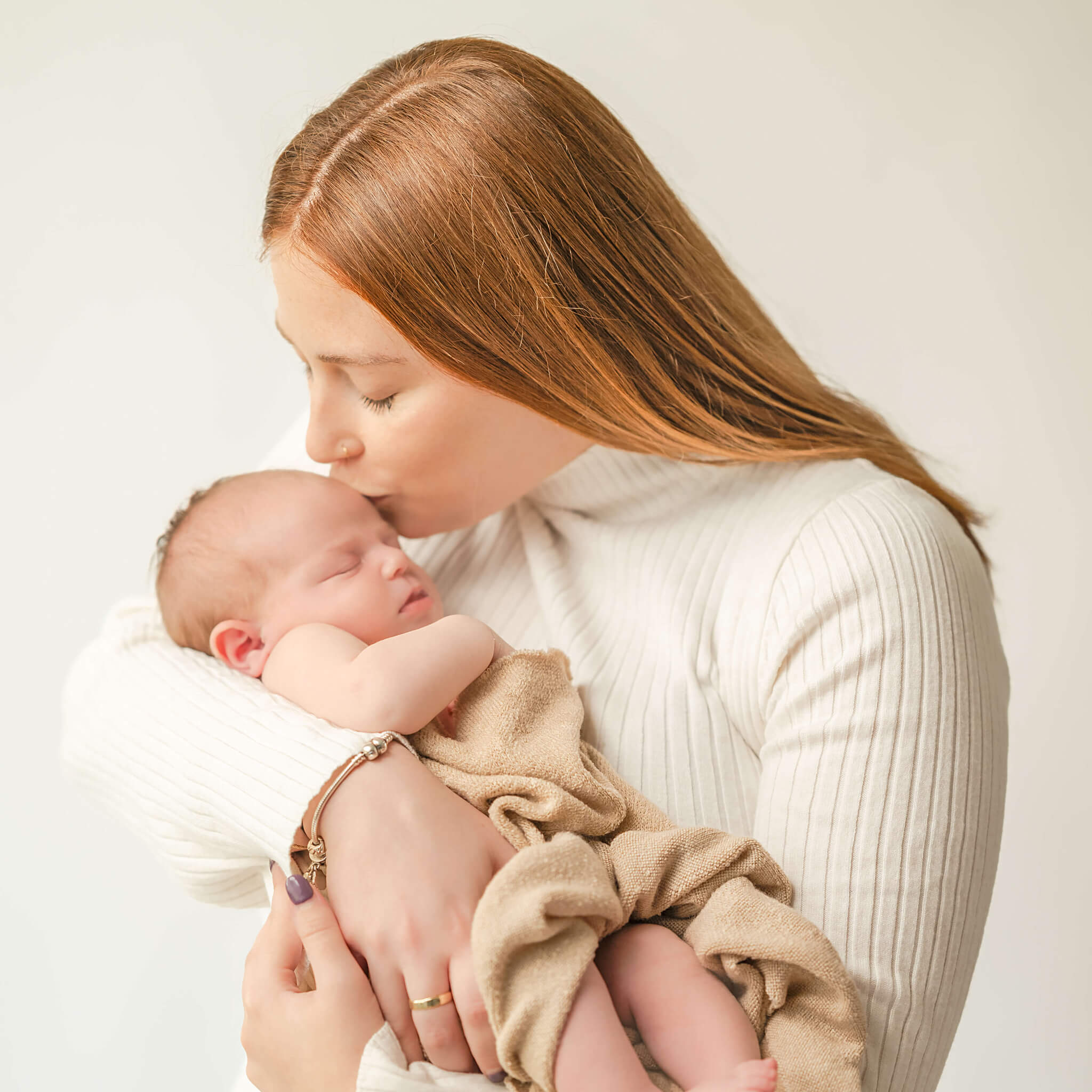 A mother in an off-white sweater dress kisses her newborn baby on the forehead. The baby is wrapped in a brown blanket.