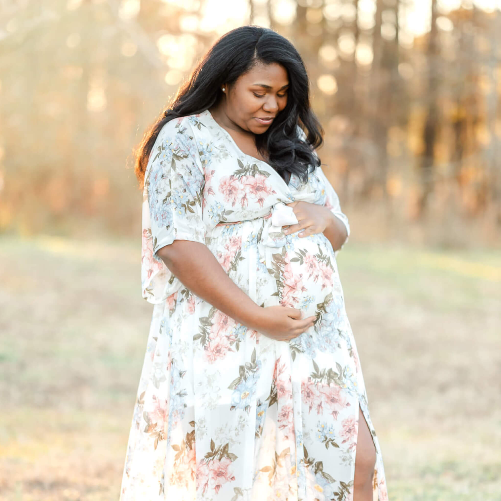 A mama-to-be in a white floral dress holds her belly and looks down. She is standing in a field, with the sun shining through the trees behind her.