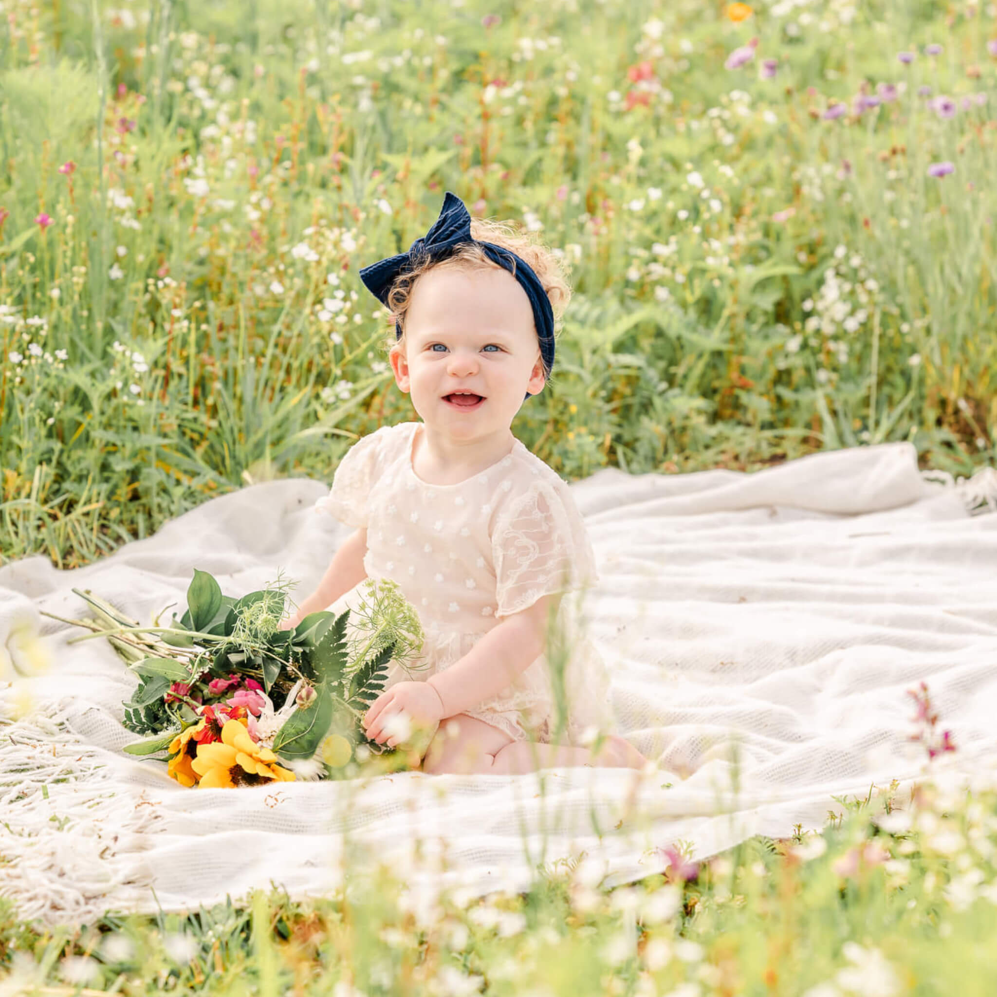 A toddler wearing a n off-white lace dress and navy blue headband sits on a white blanket in a flower field. She holds a bouquet of flowers in front of her.