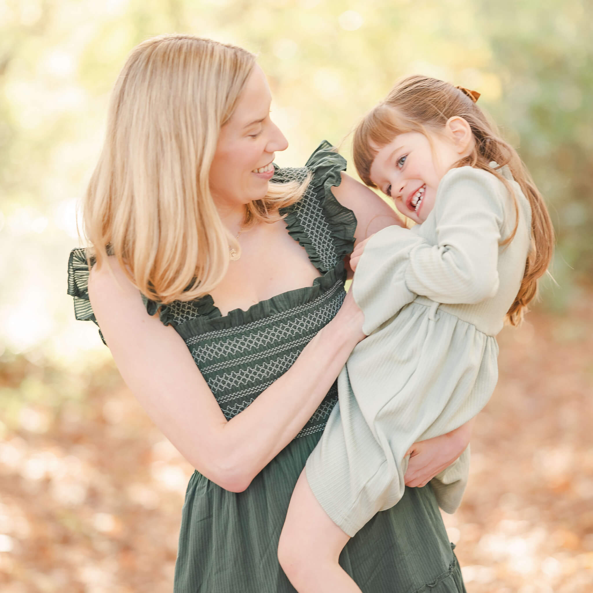 A mother with shoulder length blond hair and a green, ruffly dress, holds her toddler daughter in her arms. The little girl, who has long brown hair and is wearing a light green dress, laughs while her mom tickles her.