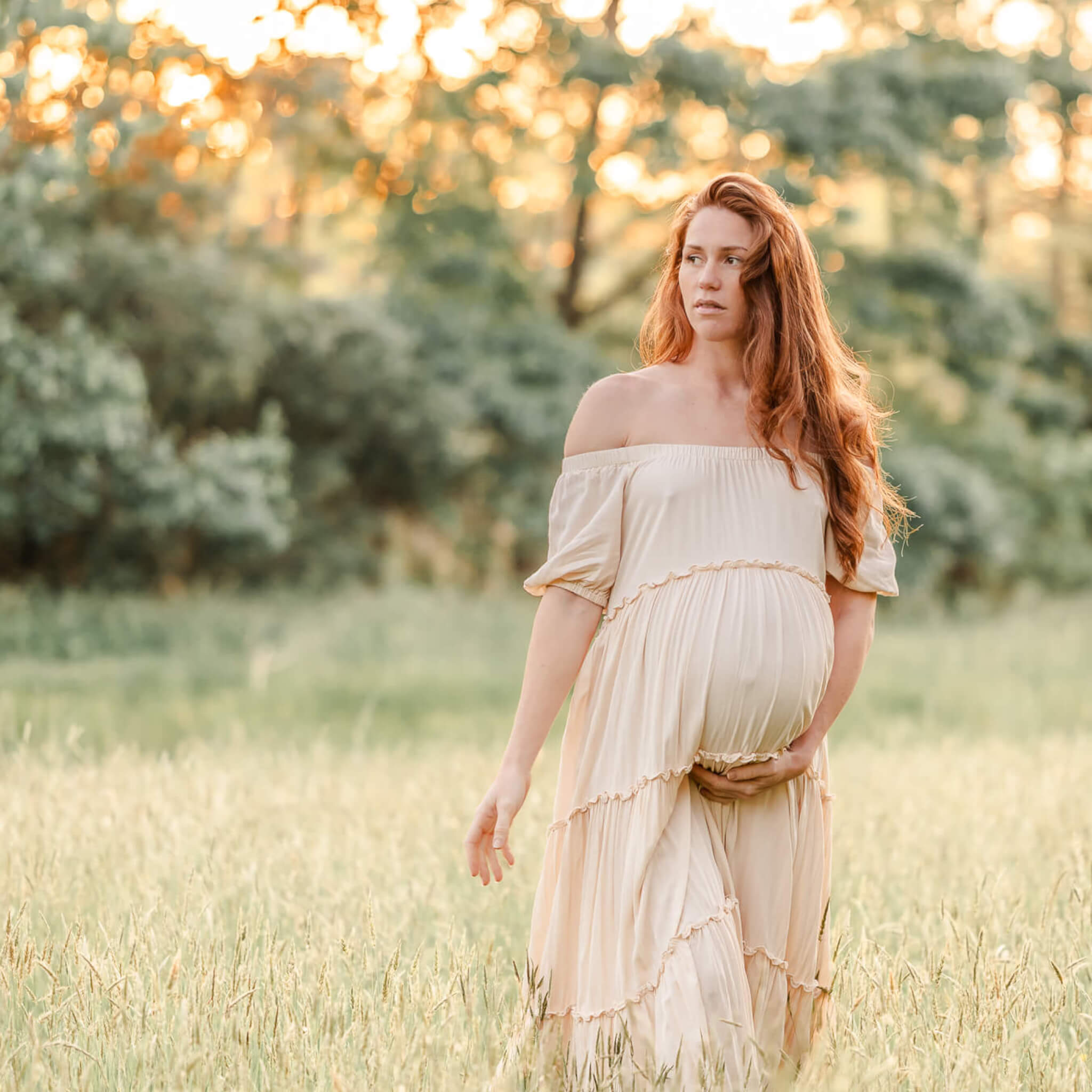 A mama-to-be, wearing an off-white flowy dress, walks through a field of tall green grass. The sun shines through the trees behind her and her long red-hair is in loose waves down her back.