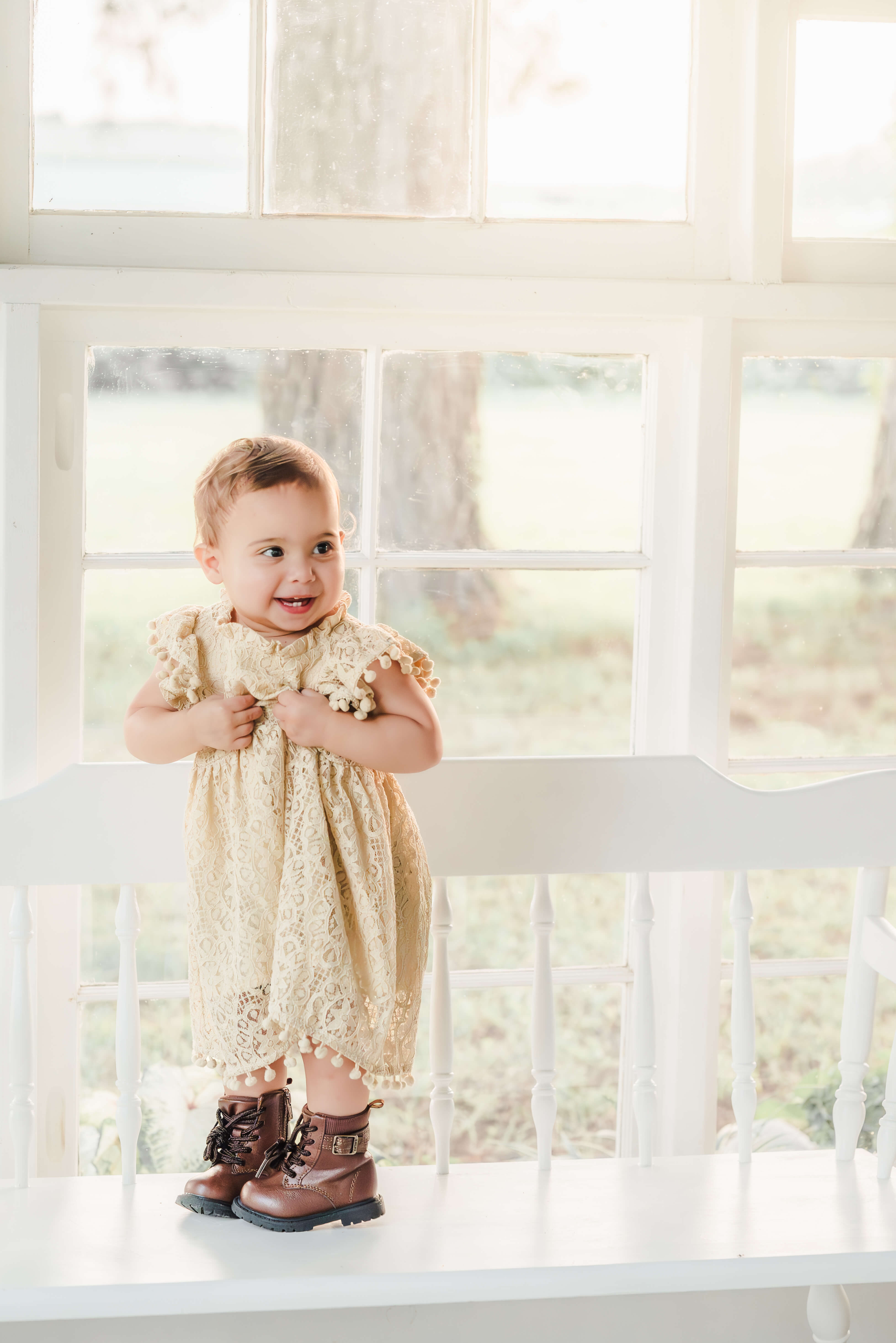 A toddler in an off-white lace dress and brown boots, stands on a white bench in a sun porch. The sun is shining through the white windows behind her. She is smiling big, showing off the few teeth she has.