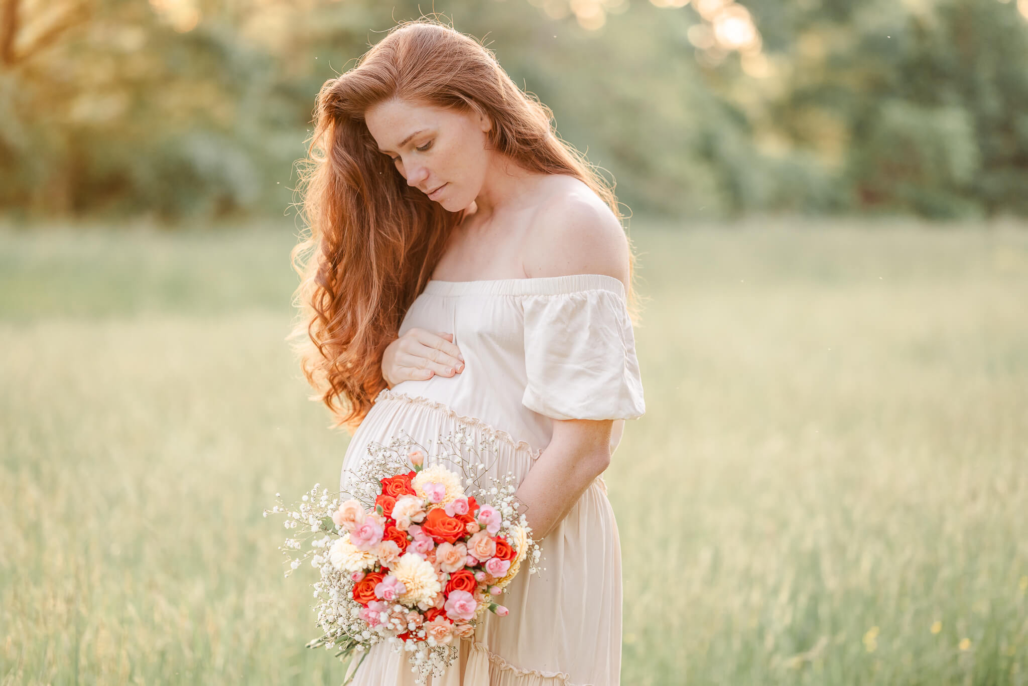 An expectant mother holds a bouquet of pink, red, and white flowers while resting her other hand on her belly. She is standing in a grassy field. The sun is shining on her long, wavy red hair. She wears a simple off-white dress.