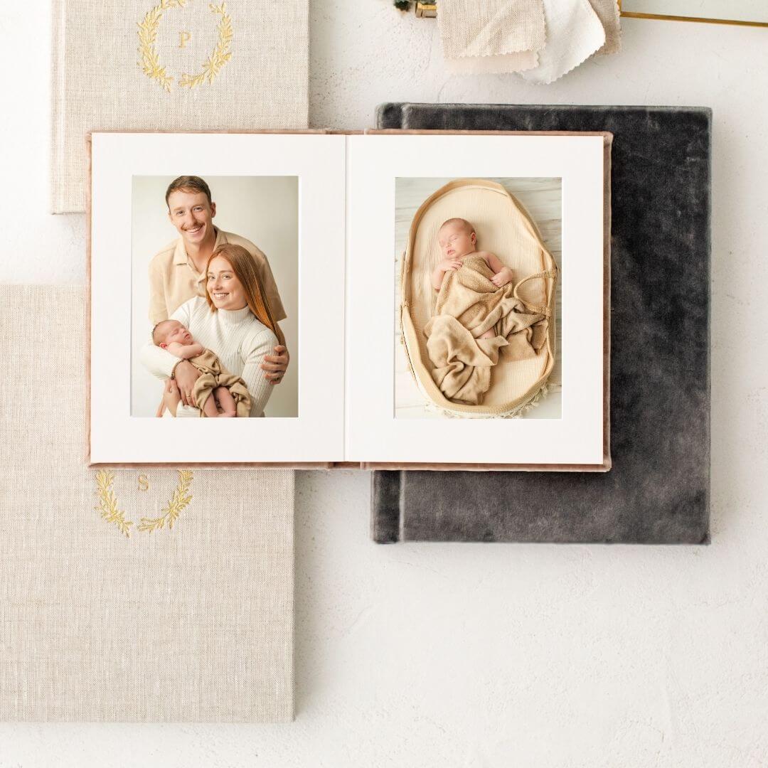 A pile of linen heirloom albums with gold embossed covers. The one on top is open and shows images from a newborn session.