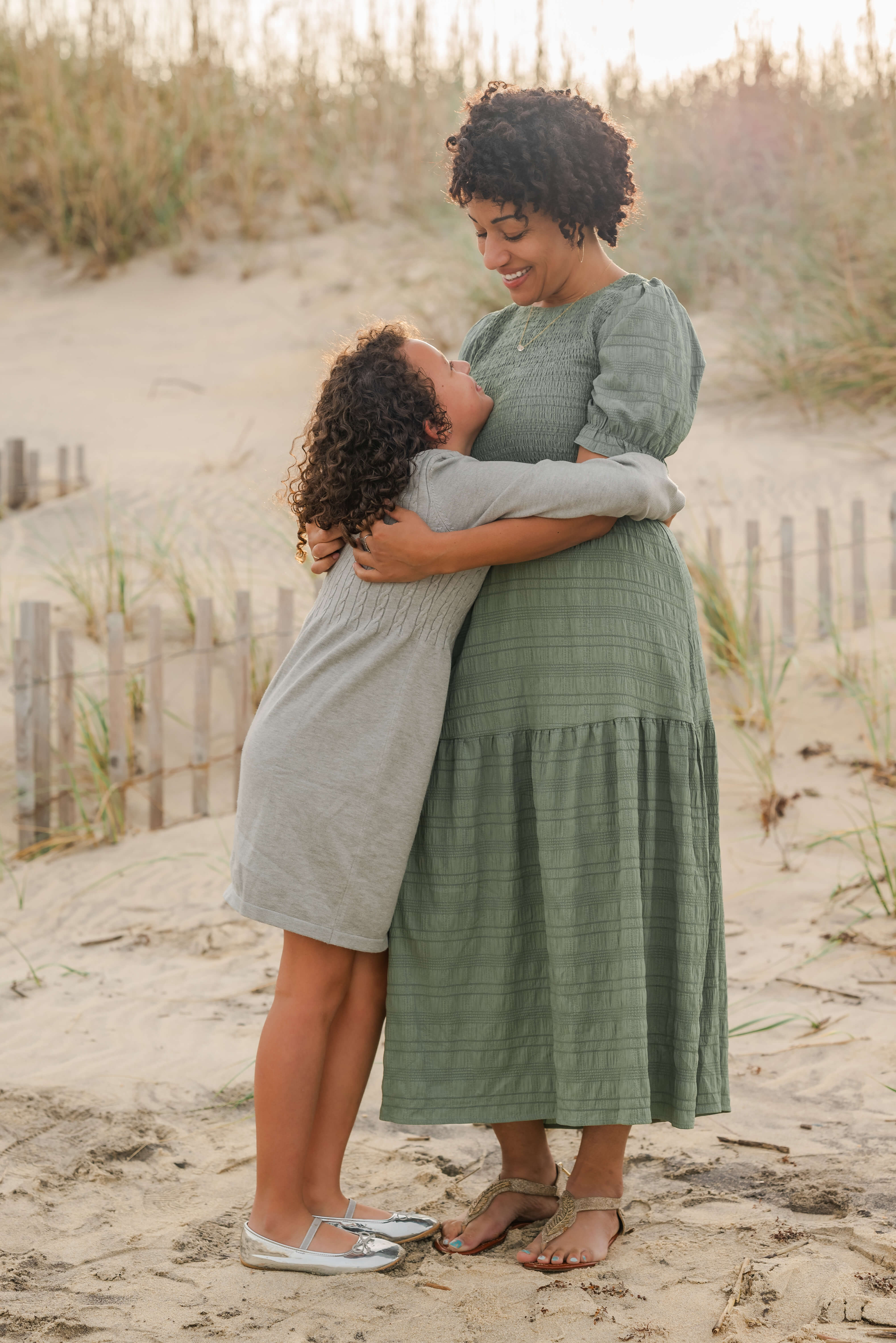 A mother in a green dress hugs her daughter who is in gray. They both look into each other's eyes while smiling. The photo is taken in front of a fence at the dunes on the beach.